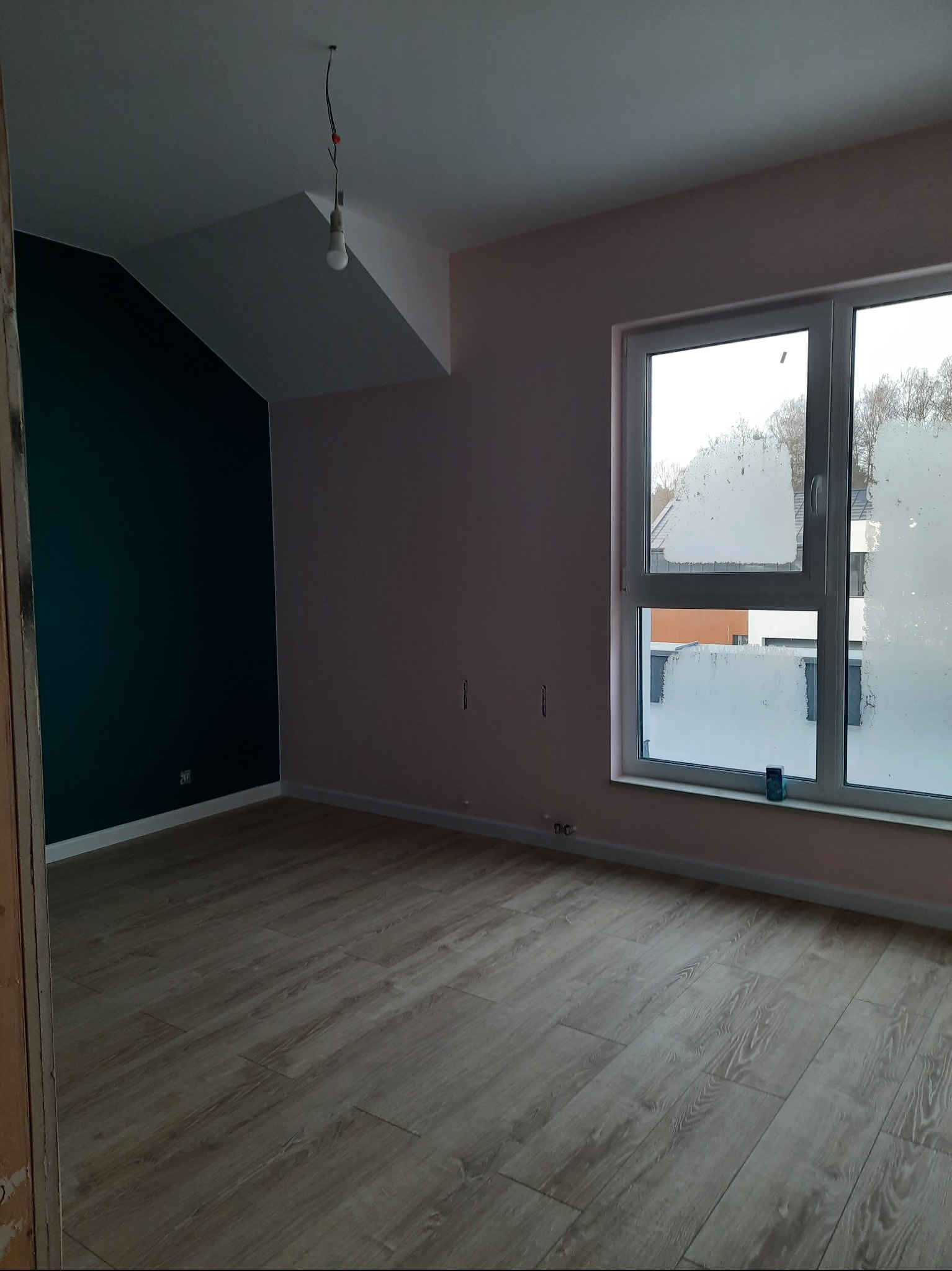 Pokój po remoncie: świeżo ułożone panele podłogowe, pomalowane ściany w odcieniach różu i zieleni, okno z widokiem na zewnątrz. Wnętrze gotowe do umeblowania.