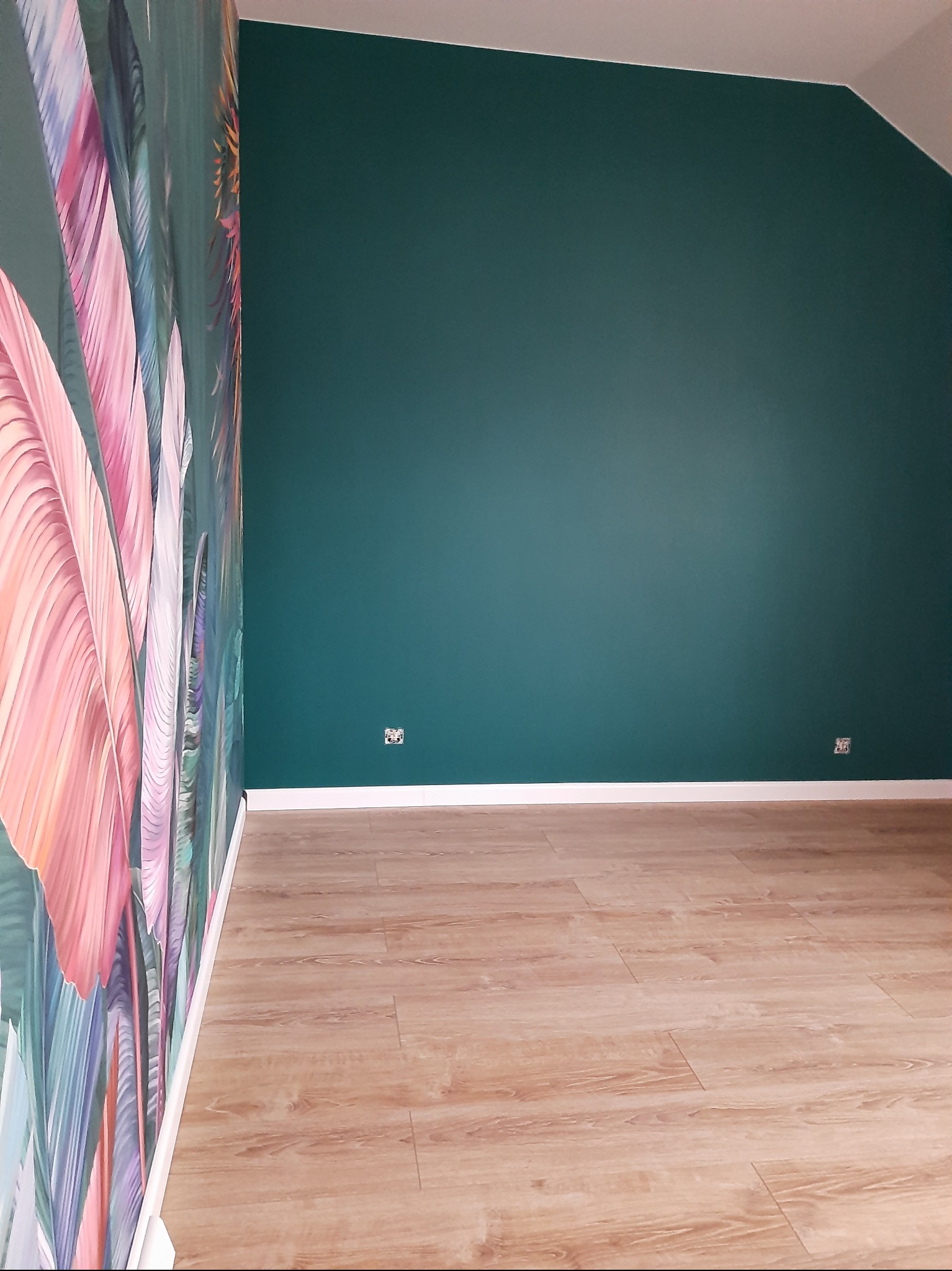 Pokój po remoncie w Łomiankach: ściana w kolorze butelkowej zieleni, wzorzysta tapeta z motywem liści i jasne panele podłogowe. Widoczne gniazdka elektryczne.