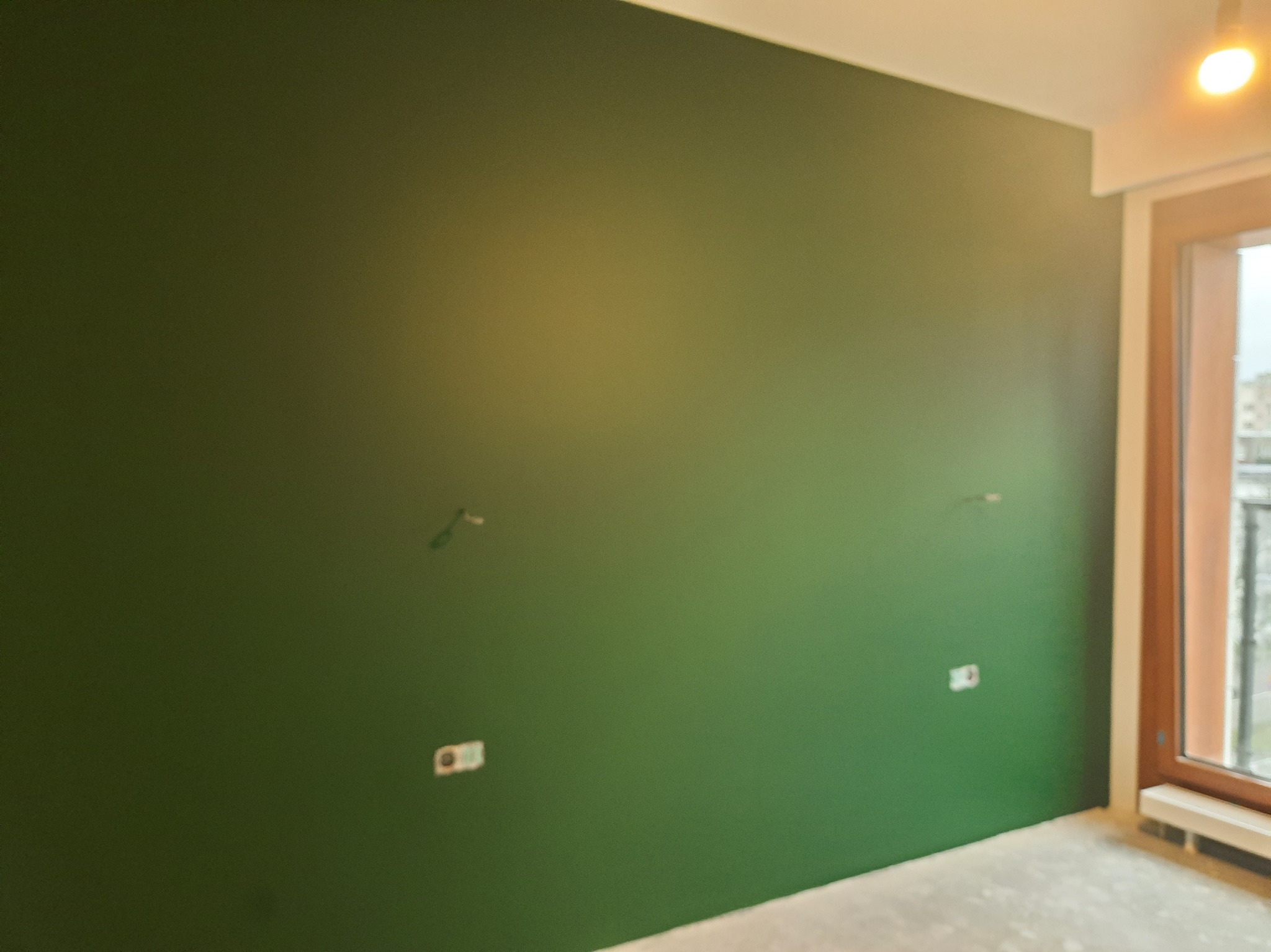 Ściana pomalowana na zielono w pokoju z oknem. Widoczne gniazdka elektryczne i niedoskonałości w wykończeniu. Świeżo pomalowane wnętrze przed dalszym wykończeniem.