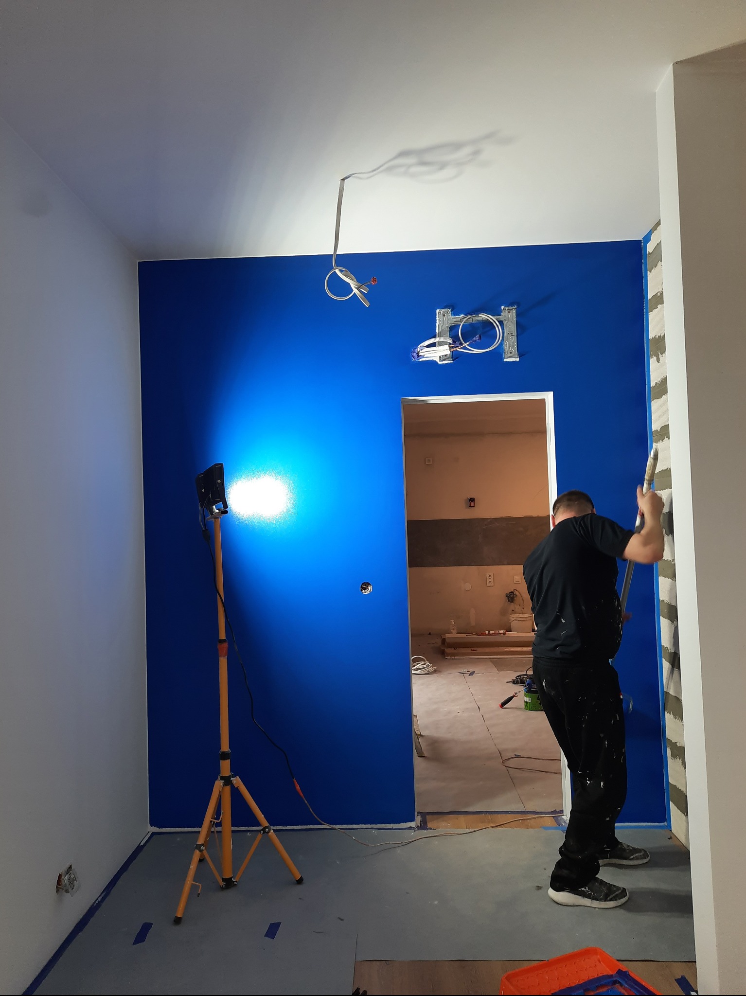 Ściana w intensywnym odcieniu niebieskiego, oświetlona lampą budowlaną, malowana przez pracownika. Widoczne przygotowania do prac wykończeniowych.