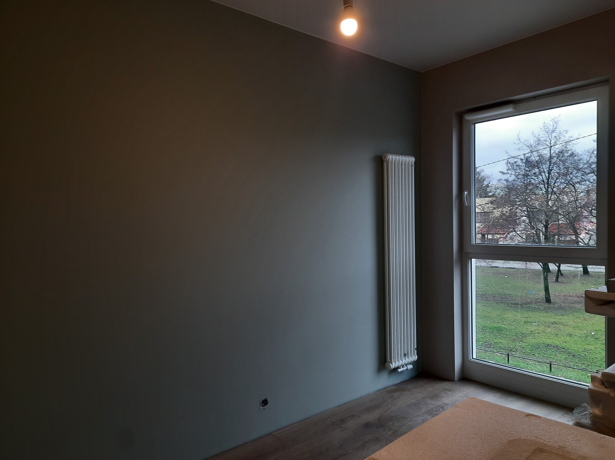 Ściana w odcieniach szarości z białym grzejnikiem i oknem z widokiem na zewnątrz. Podłoga wykończona panelami. Minimalistyczne wnętrze po remoncie.