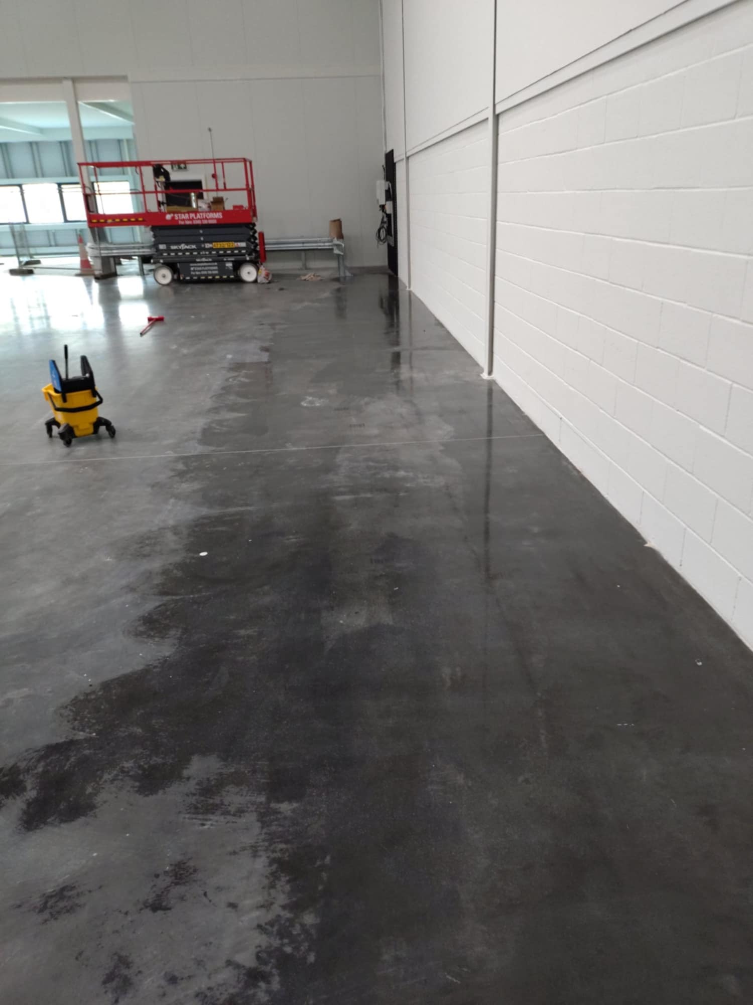 Wnętrze hali po malowaniu posadzki żywicą epoksydową. Widoczny sprzęt do czyszczenia, podnośnik i świeżo pomalowana, błyszcząca posadzka w szarym kolorze.