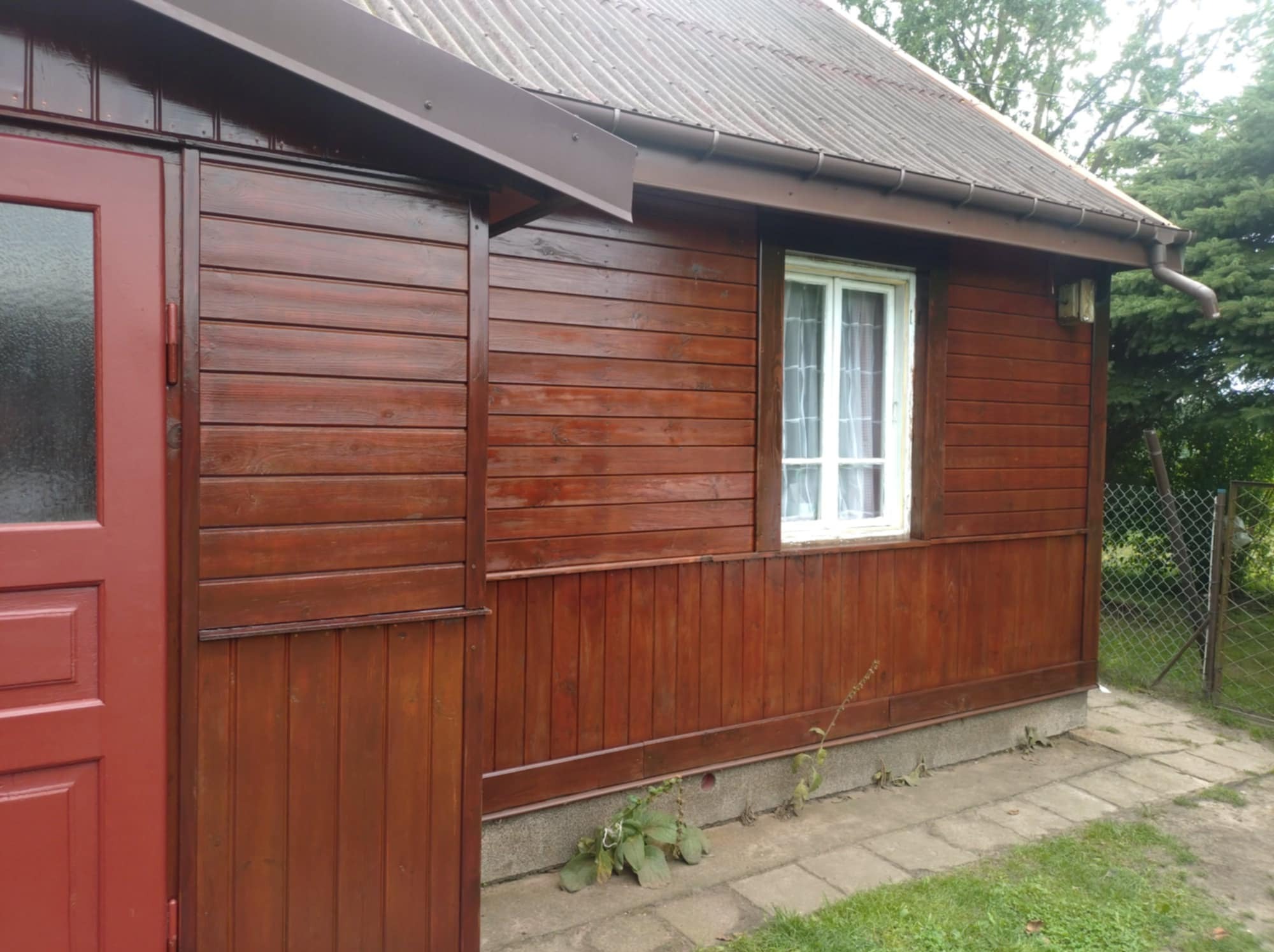 Pomazany na brązowo drewniany budynek z białym oknem i brązowymi rynnami, widziany z boku, z fragmentem chodnika i zieleni w tle.