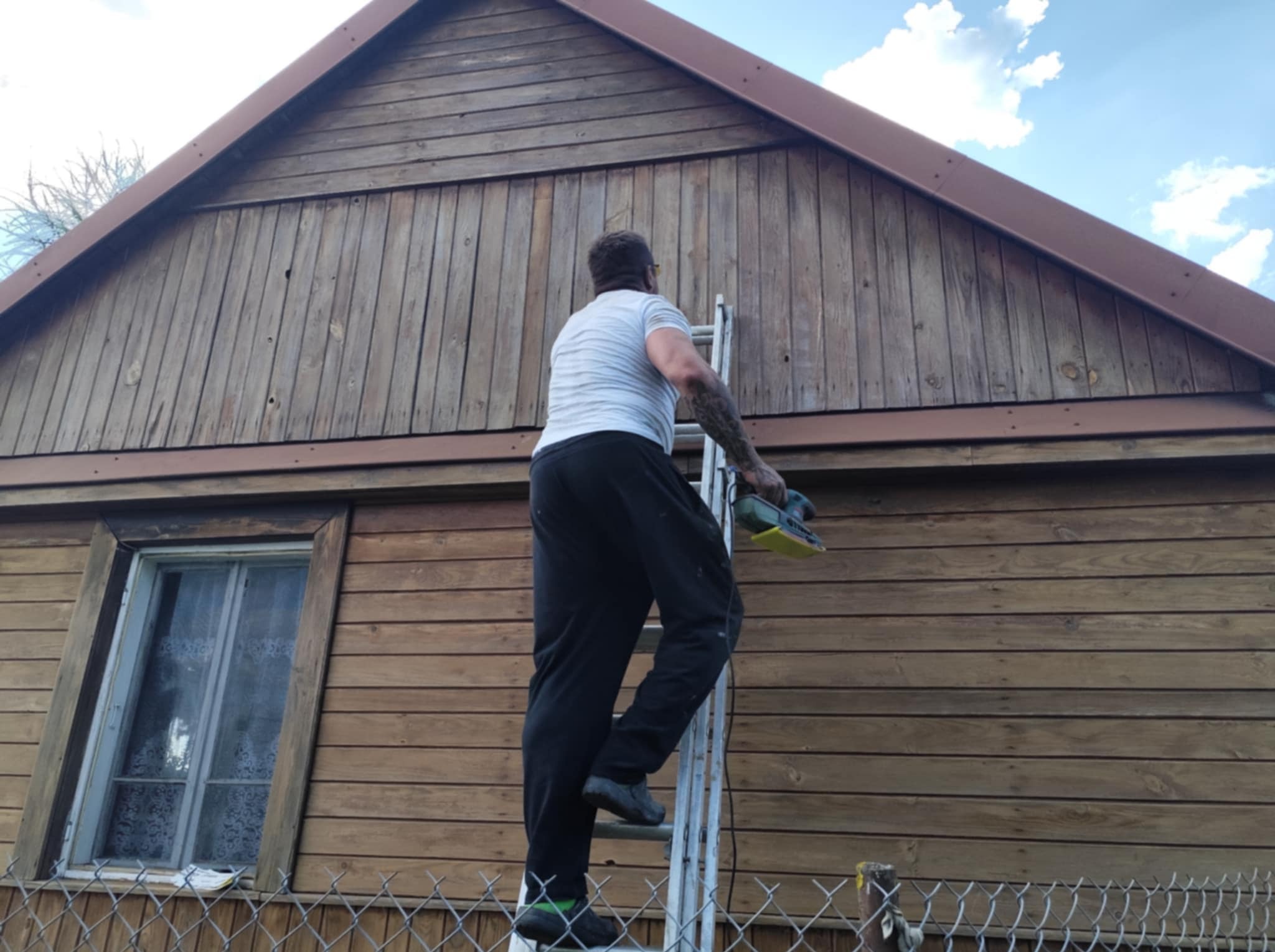 Mężczyzna na drabinie szlifuje drewnianą elewację domu przed malowaniem. Widoczne okno i fragment ogrodzenia. Prace renowacyjne elewacji.
