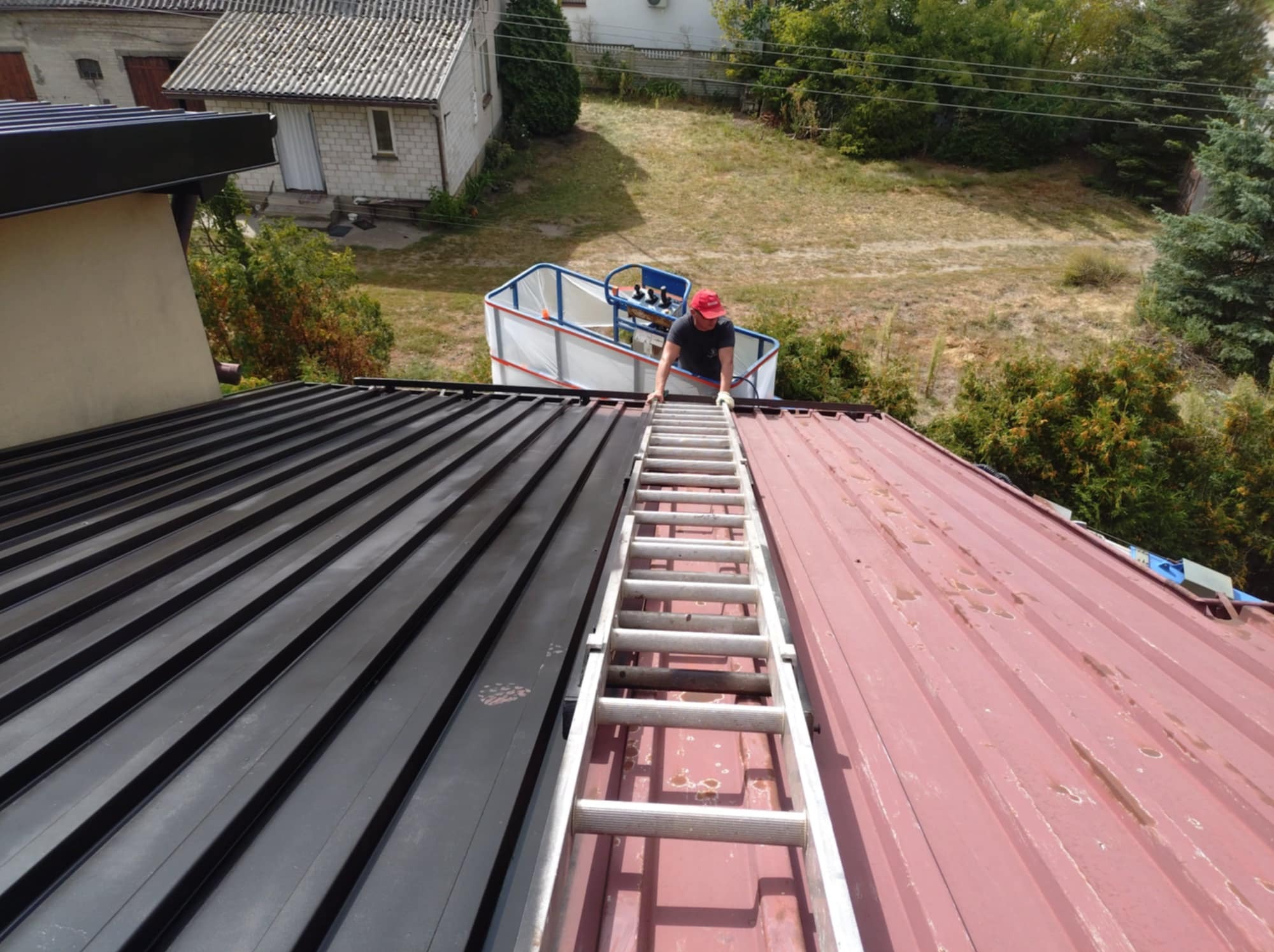 Malowanie dachu z blachy trapezowej na czarno. Widoczna drabina i pracownik w czerwonej czapce. Połowa dachu pomalowana, druga w kolorze czerwonym.