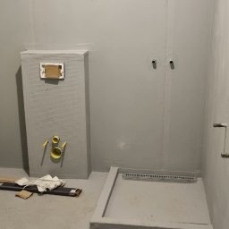 LG Jaworzno - Szara łazienka w trakcie prac wykończeniowych. Widoczny brodzik z odpływem liniowym, zabudowa stelaża WC z płyt karton-gips i instalacje. Surowy stan, przygotowanie do dalszych prac.