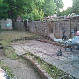 LG Jaworzno - Zaniedbany ogród z drewnianym płotem, patio z płyt, narzędzia ogrodnicze na stole i zarośnięte ścieżki. Widoczny domek narzędziowy i zieleń drzew.