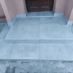 LG Jaworzno - Wykończone płytkami schody wejściowe do domu, z ciemnymi drzwiami w tle. Jasnoszare płytki ułożone równo, z widocznymi fugami. Detal wykończenia.