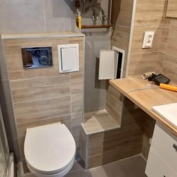 LG Jaworzno - Wykończona łazienka z podwieszaną toaletą, zabudową z płytek imitujących drewno i szafką z drewnianym blatem. Widoczny bojler i otwarta klapka rewizyjna.