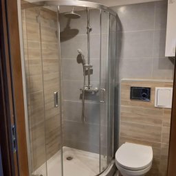 LG Jaworzno - Nowoczesna kabina prysznicowa z zaokrąglonymi drzwiami i toaleta w wykończonej łazience z szarymi i drewnopodobnymi płytkami. Widok zza futryny drzwi.