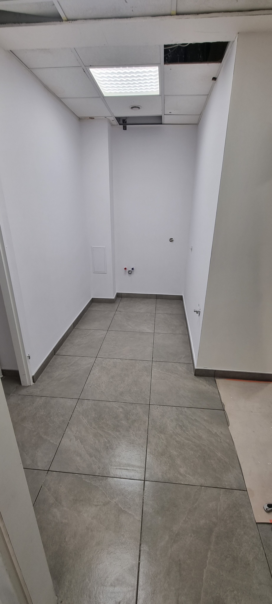 Wnętrze po remoncie: szare płytki podłogowe, białe ściany z widocznymi instalacjami. Sufit podwieszany z oświetleniem i fragmentem odsłoniętej konstrukcji.