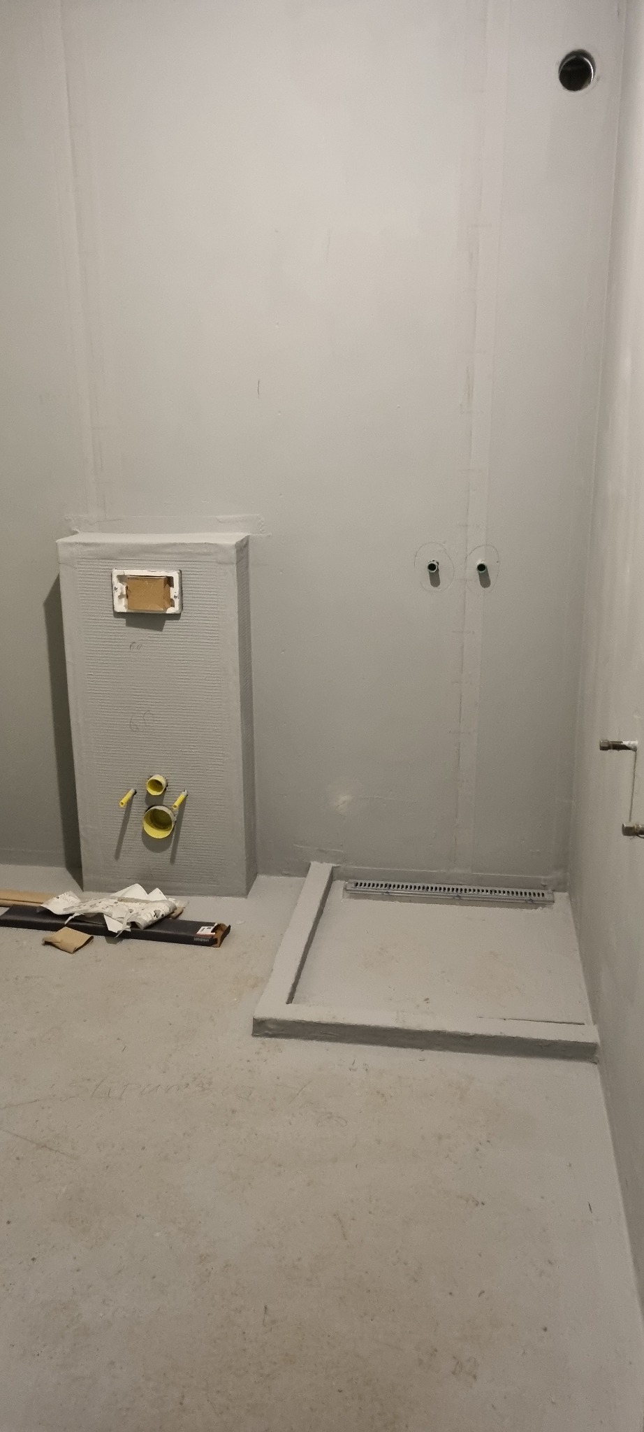 Szara łazienka w trakcie prac wykończeniowych. Widoczny brodzik z odpływem liniowym, zabudowa stelaża WC z płyt karton-gips i instalacje. Surowy stan, przygotowanie do dalszych prac.