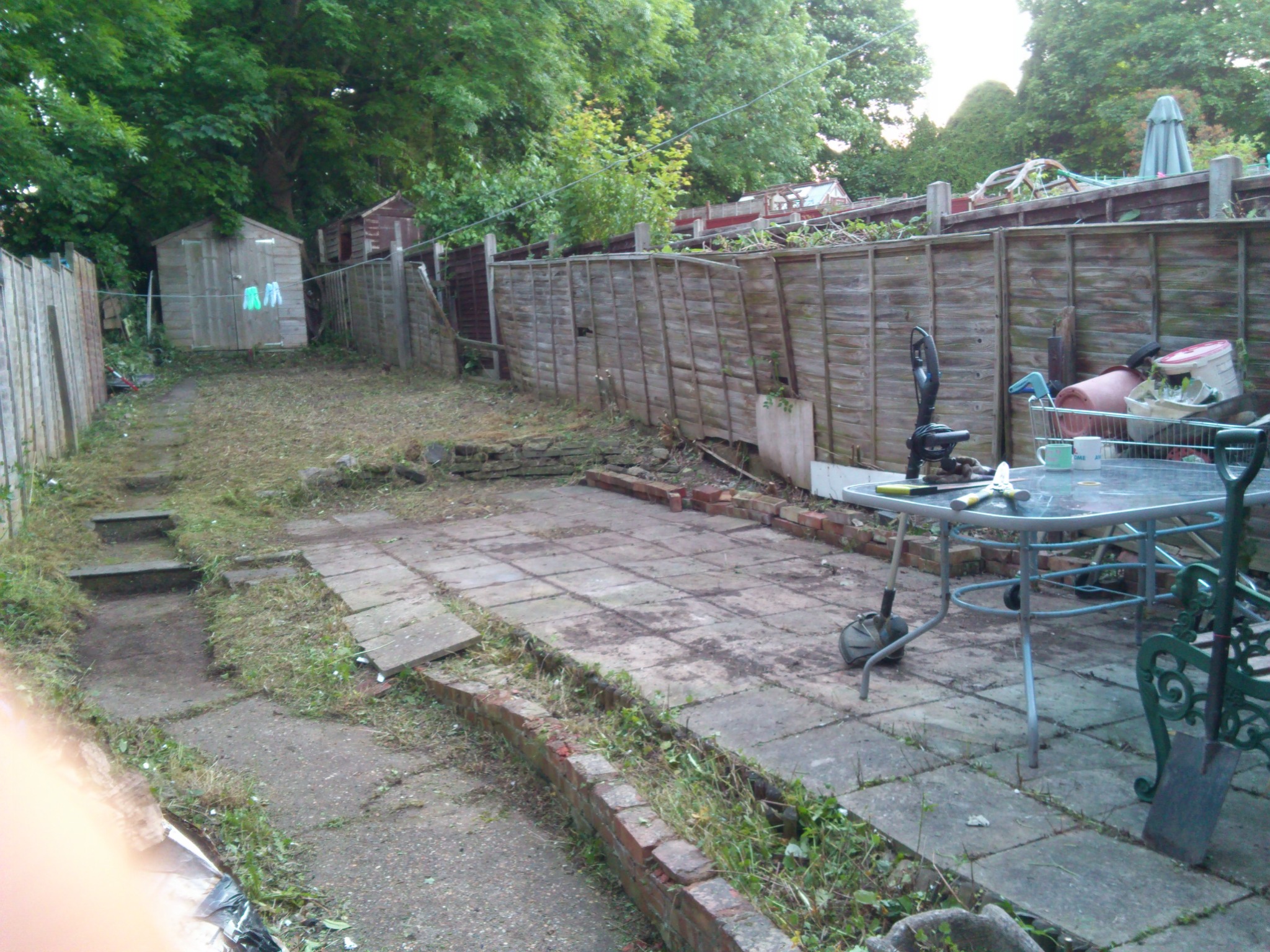 Zaniedbany ogród z drewnianym płotem, patio z płyt, narzędzia ogrodnicze na stole i zarośnięte ścieżki. Widoczny domek narzędziowy i zieleń drzew.