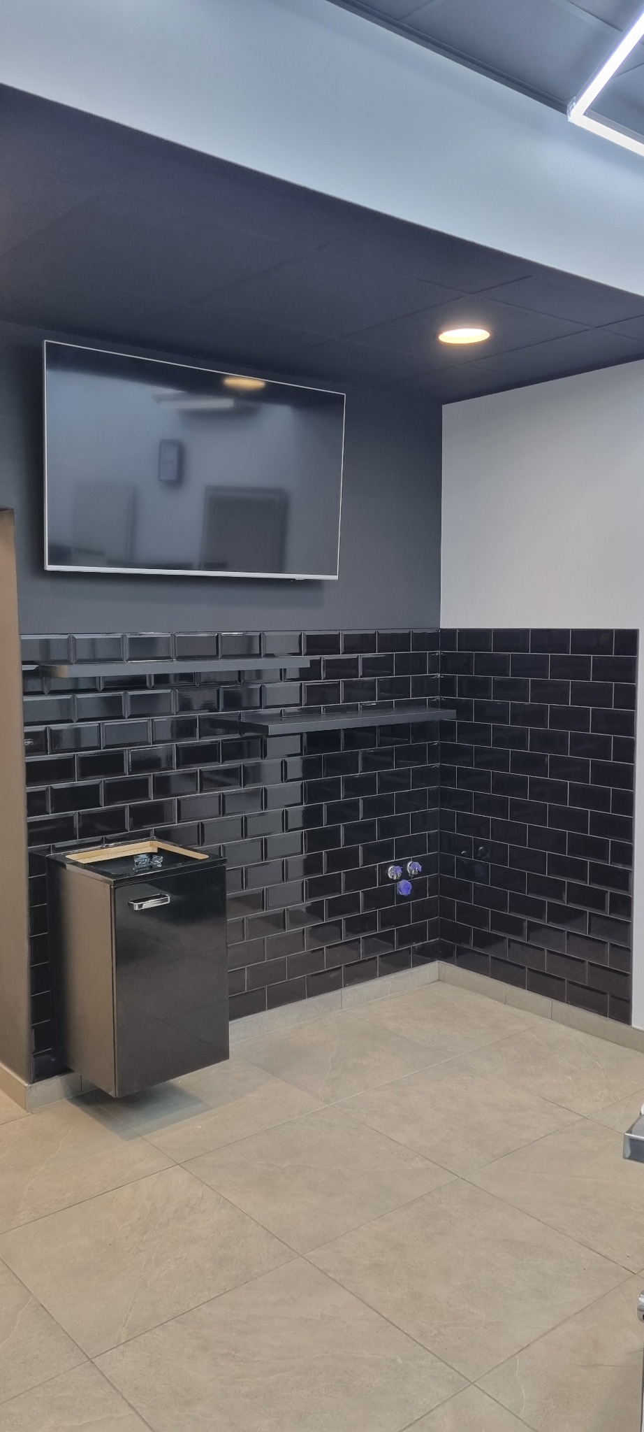 Nowoczesna łazienka z czarnymi, błyszczącymi płytkami typu metro na ścianach, wiszącą szafką pod umywalkę i dużym telewizorem na ścianie. Jasna podłoga z płytek.