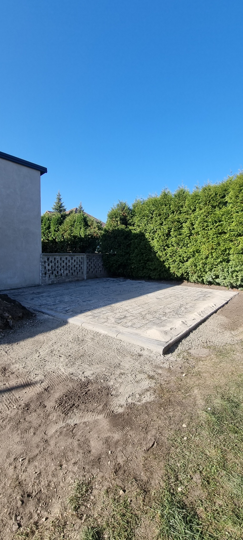 Przygotowany teren pod kostkę brukową z obrzeżami, widok z góry, w tle budynek i tuje. Teren wyrównany, gotowy do dalszych prac brukarskich.