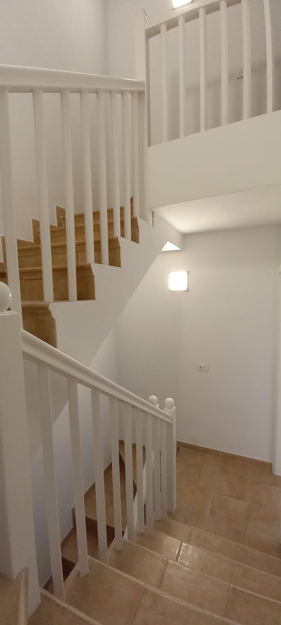 Jasne schody z białymi balustradami i beżowymi płytkami podłogowymi. Widok z góry na dół, oświetlenie punktowe na ścianie.