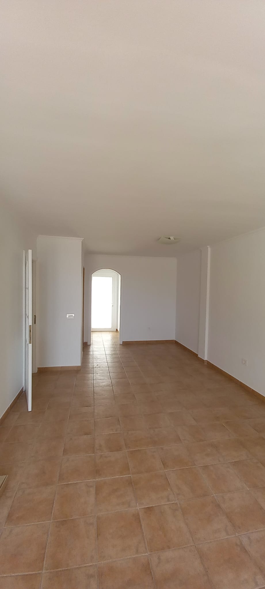 Jasne, przestronne wnętrze po remoncie z terakotą na podłodze. Świeżo pomalowane ściany na biało, widoczne drzwi i przejście do kolejnego pomieszczenia.