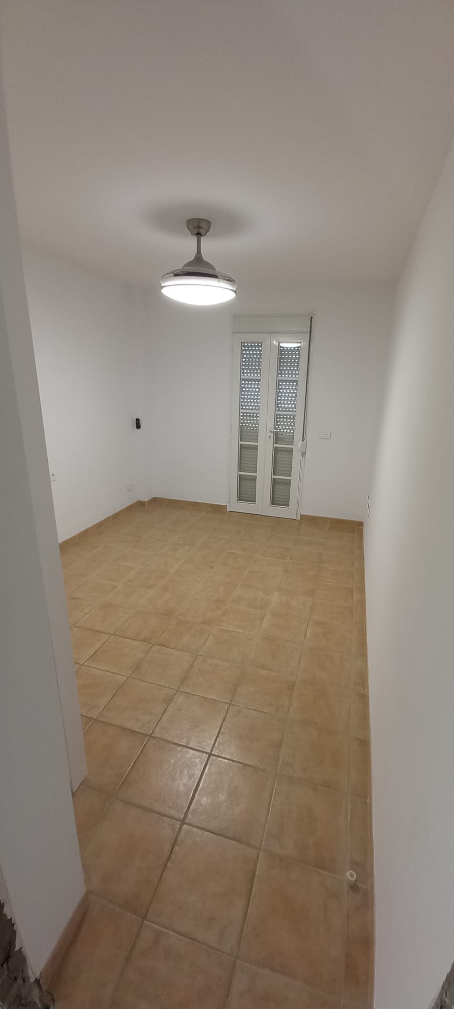 Jasne wnętrze po odświeżeniu: białe ściany, brązowa podłoga z płytek, nowoczesna lampa sufitowa i białe, przeszklone drzwi balkonowe. Minimalistyczny design.