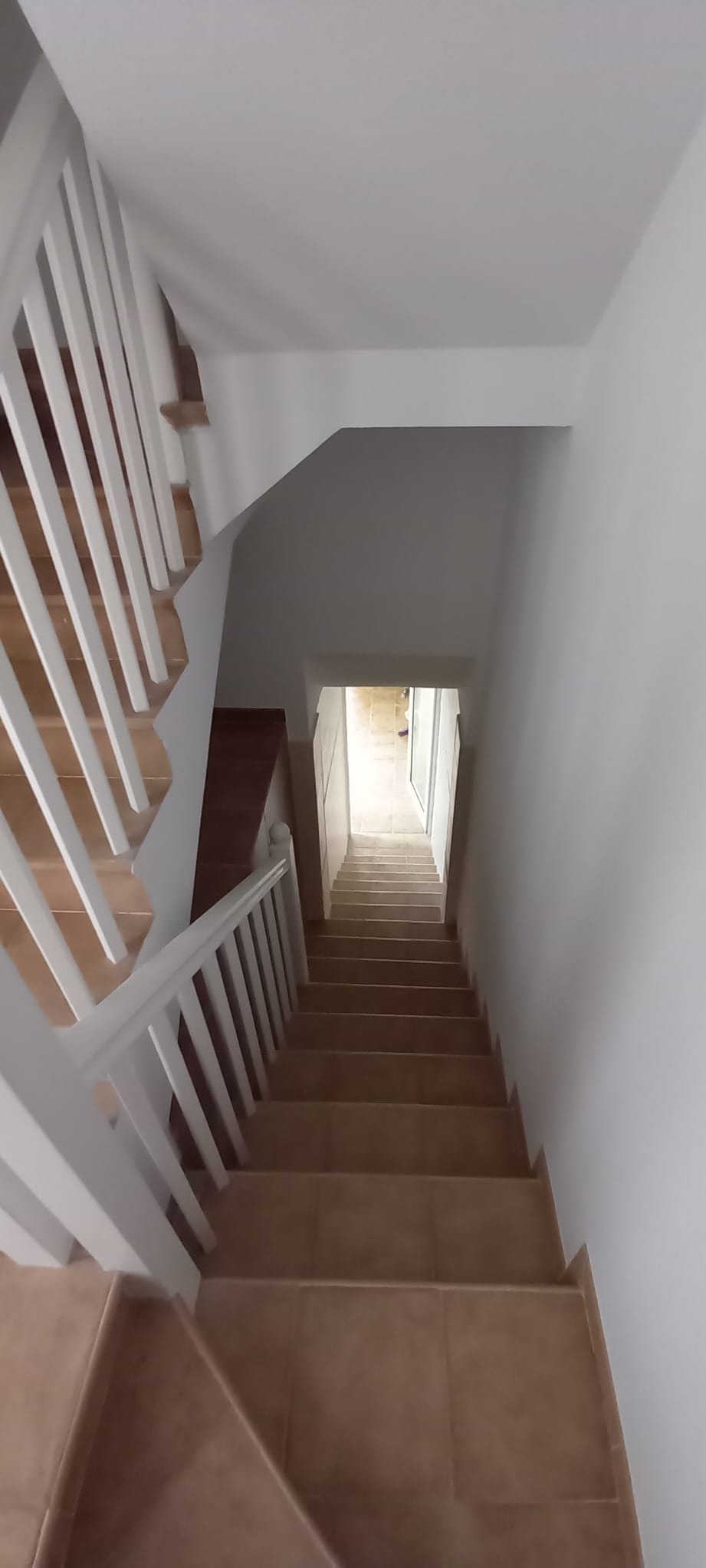 Klatka schodowa z brązowymi płytkami i białymi balustradami, widziana z góry. Ściany pomalowane na biało, wpadające światło dzienne z dołu schodów.