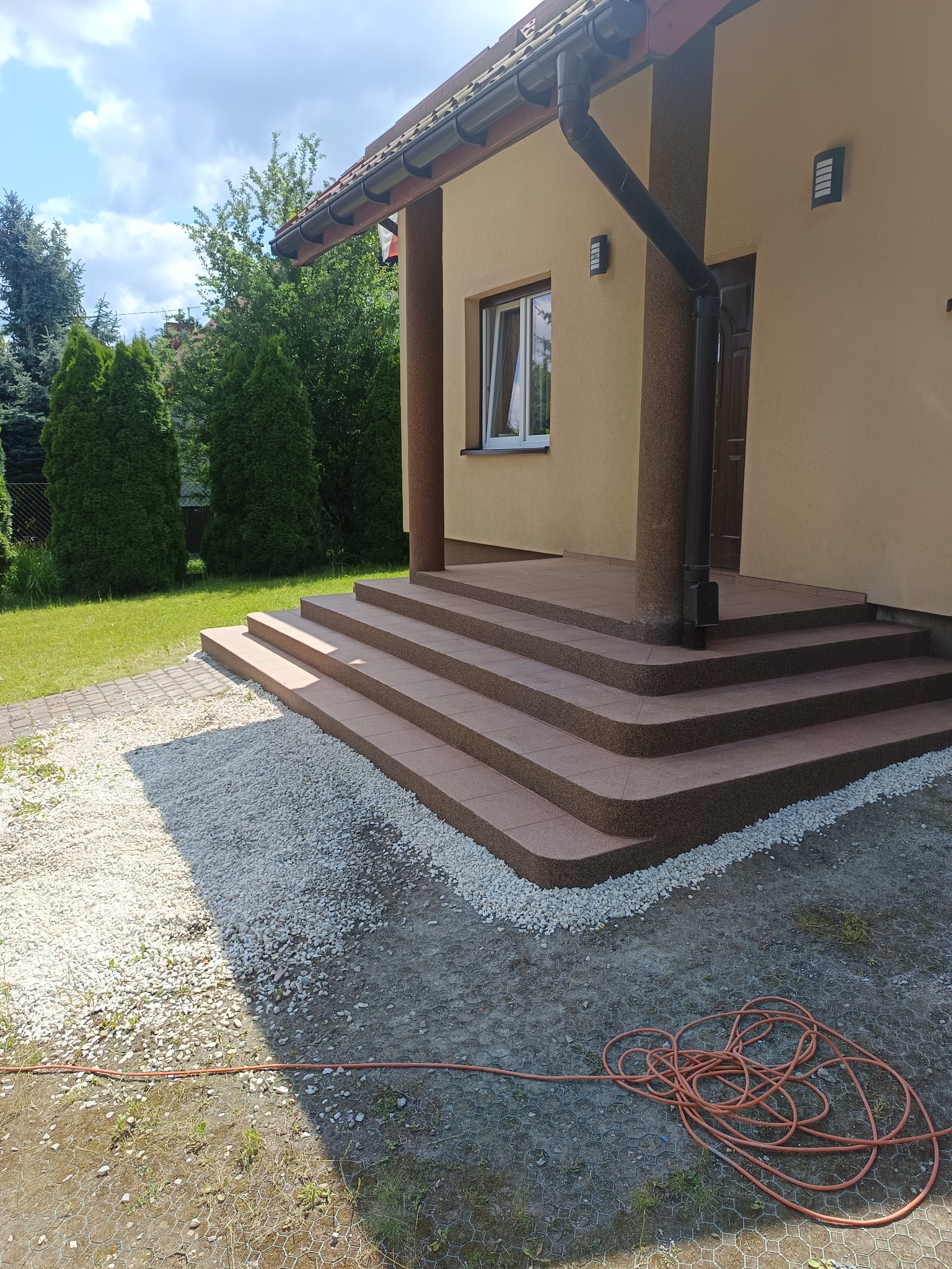 Wykończone schody wejściowe do domu z brązowych płytek, otoczone białym żwirem. Widoczny fragment elewacji w ciepłym odcieniu beżu i zielony trawnik w tle.