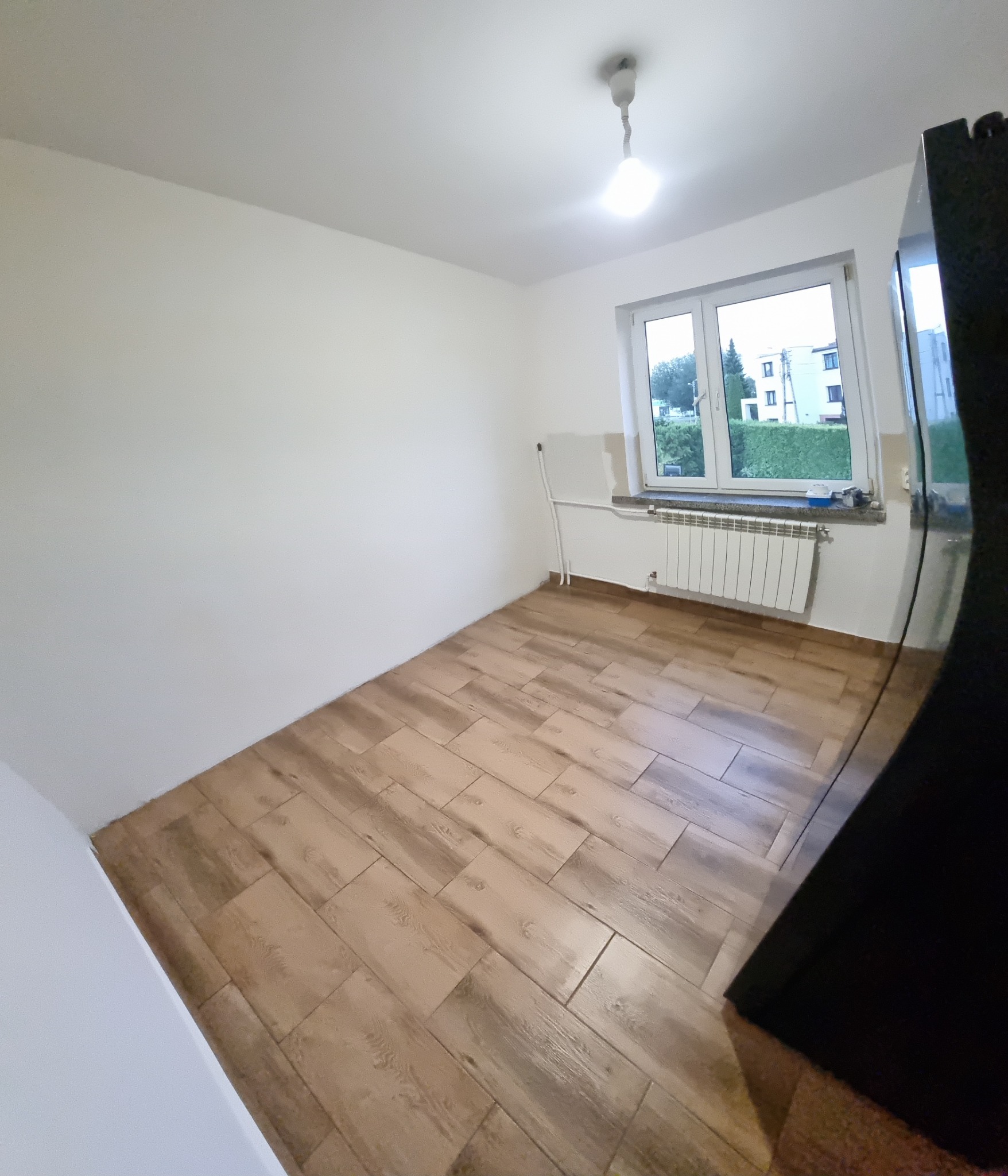 Ukończona podłoga z płytek imitujących drewno w pokoju w Jaworznie. Ściany pomalowane na biało, widoczny grzejnik pod oknem. Ujęcie z narożnika pomieszczenia.