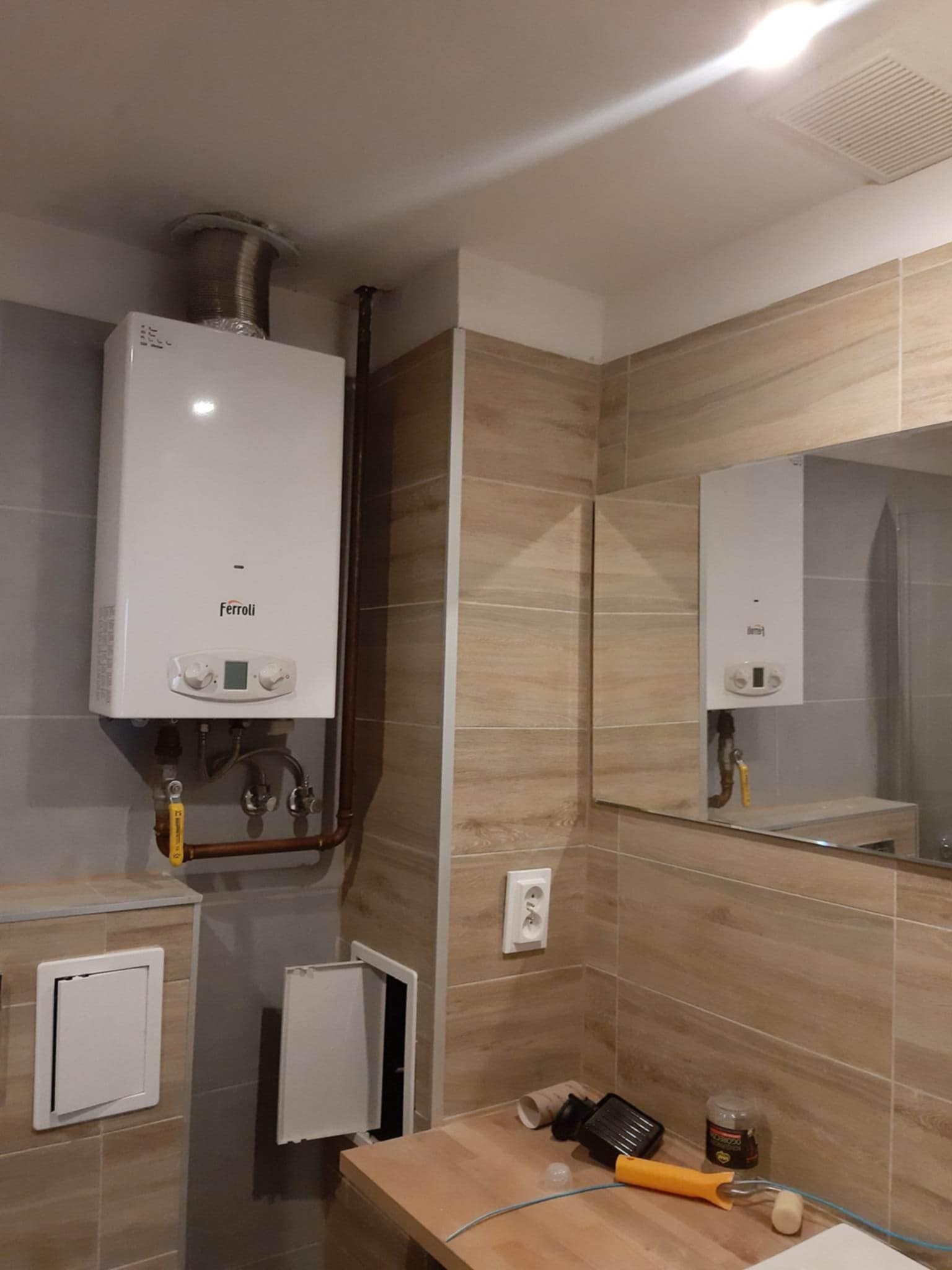 Biały piec gazowy Ferroli zamontowany w łazience z beżowymi płytkami, widoczne rury miedziane i narzędzia na blacie. Odbicie pieca w lustrze.