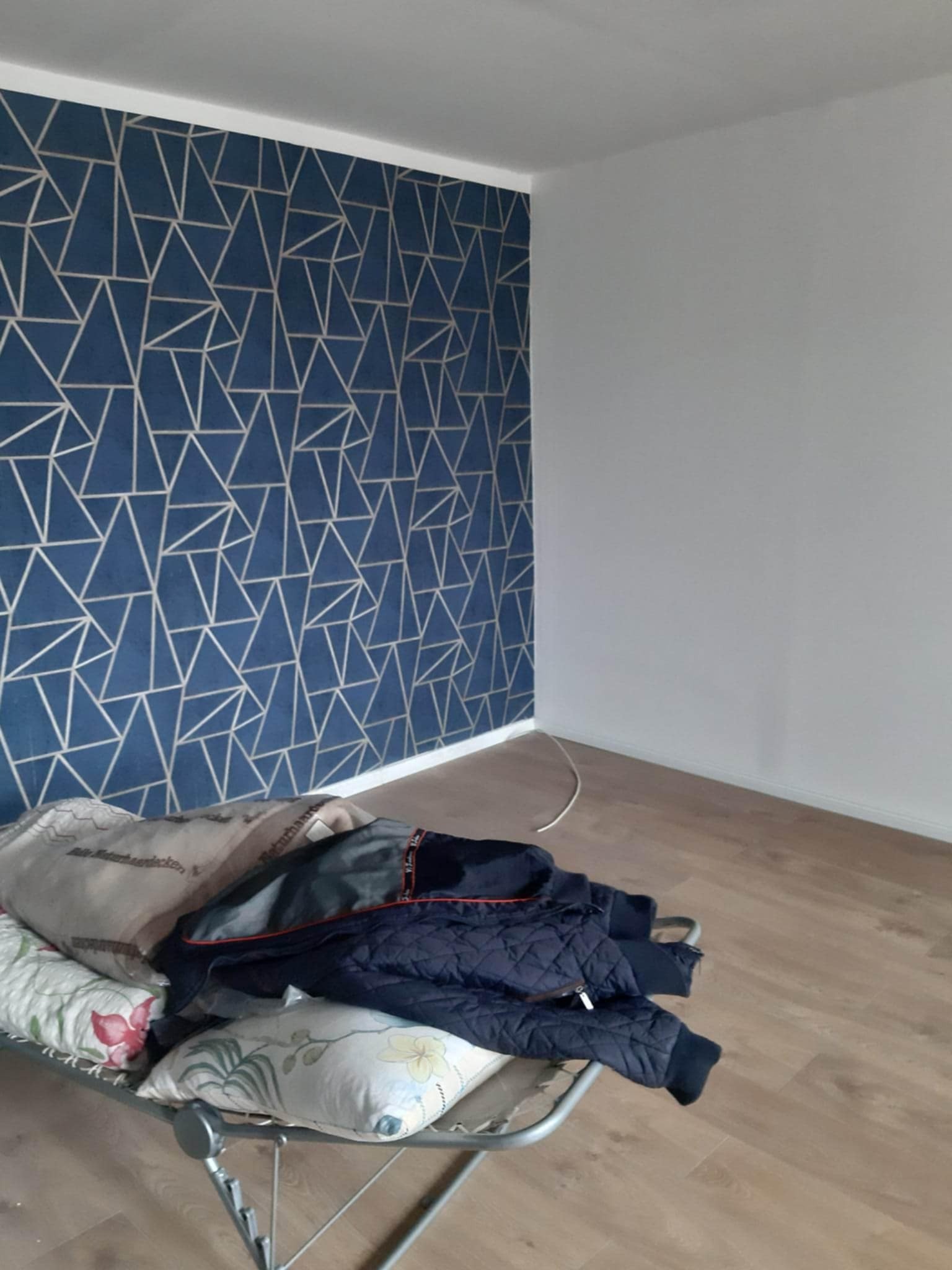 Pokój po remoncie: ściana z geometrycznym wzorem w odcieniach niebieskiego i złota, sąsiadująca z białą ścianą. Na podłodze leżą rzeczy przykryte poduszkami.