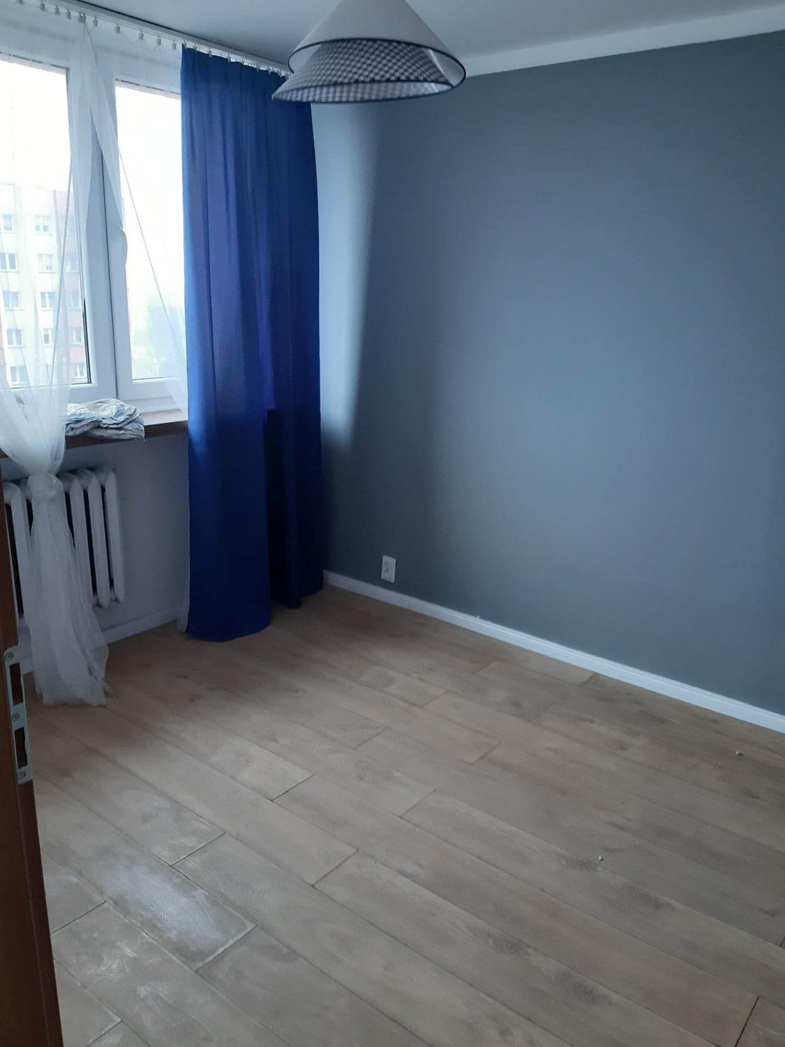 Pomalowany na szaro pokój z drewnianą podłogą, białym grzejnikiem pod oknem z firankami i niebieskimi zasłonami. Widoczna lampa sufitowa.