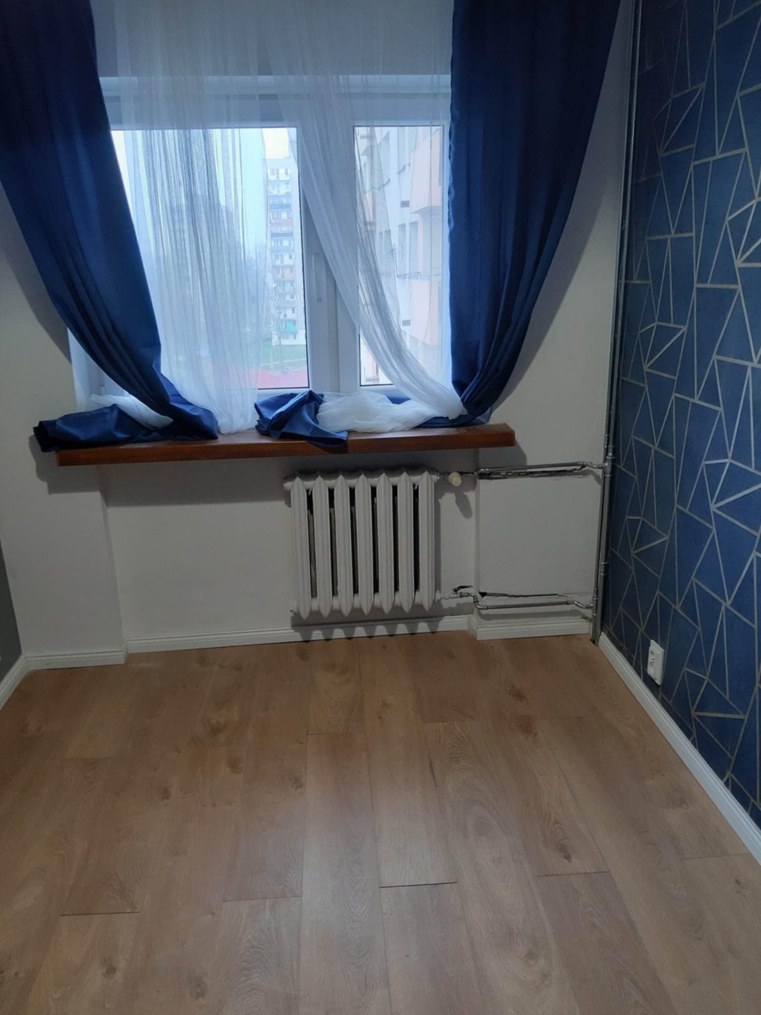 Pokój po remoncie: drewniana podłoga, biały grzejnik pod oknem z niebieskimi zasłonami, ściana z geometrycznym wzorem w odcieniach niebieskiego i złota.