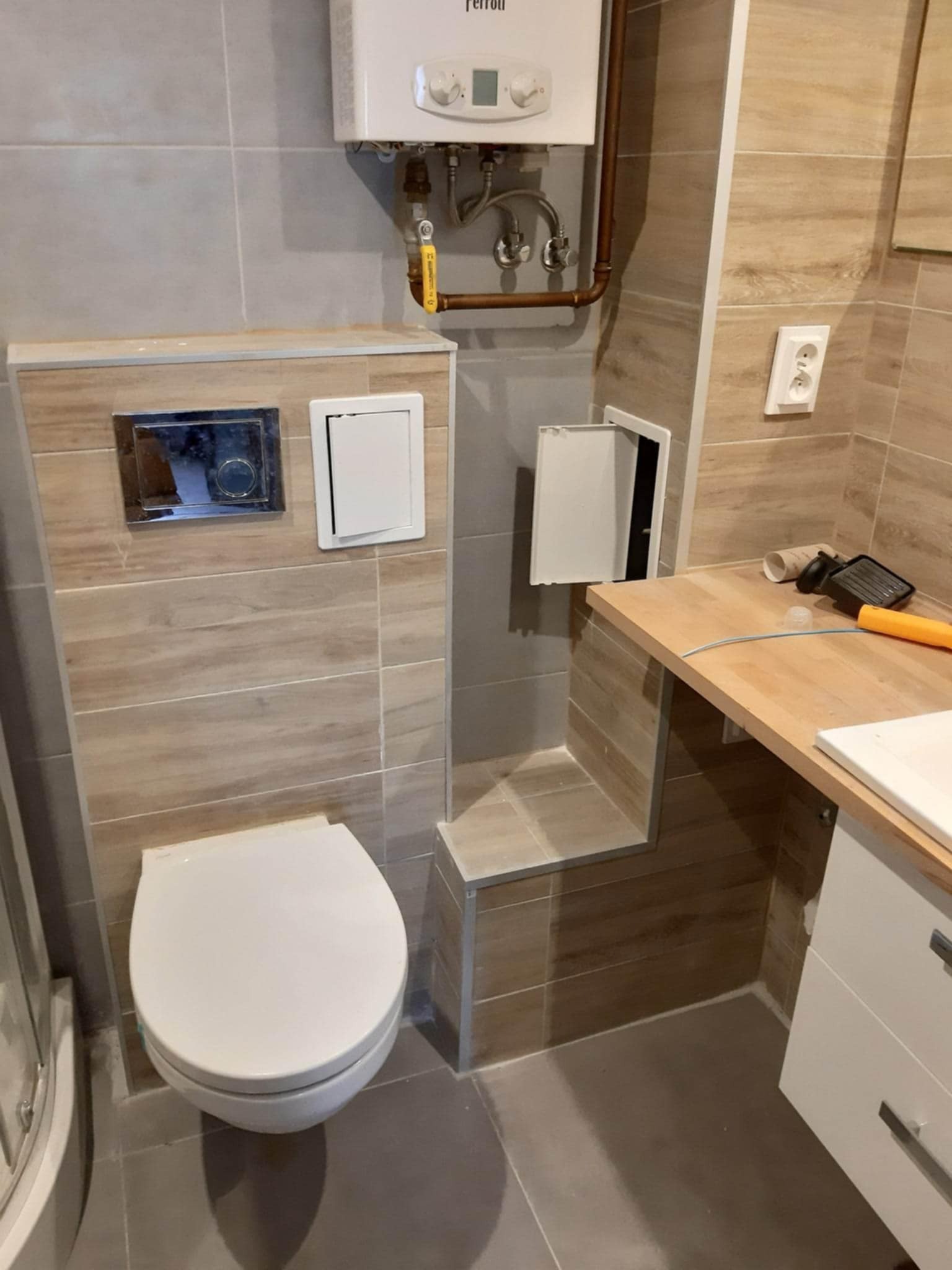 Wykończona łazienka z podwieszaną toaletą, zabudową z płytek imitujących drewno i szafką z drewnianym blatem. Widoczny bojler i otwarta klapka rewizyjna.