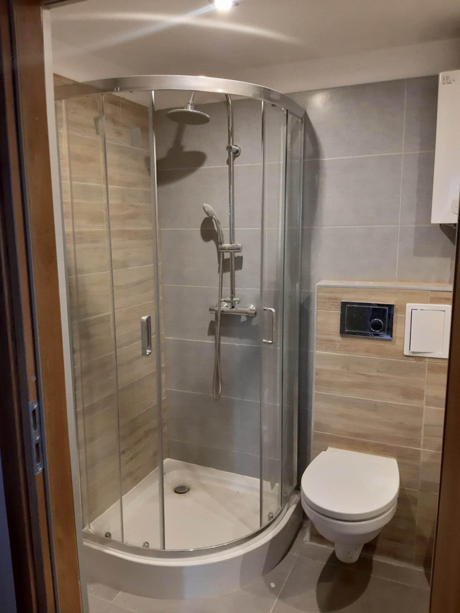 Nowoczesna kabina prysznicowa z zaokrąglonymi drzwiami i toaleta w wykończonej łazience z szarymi i drewnopodobnymi płytkami. Widok zza futryny drzwi.