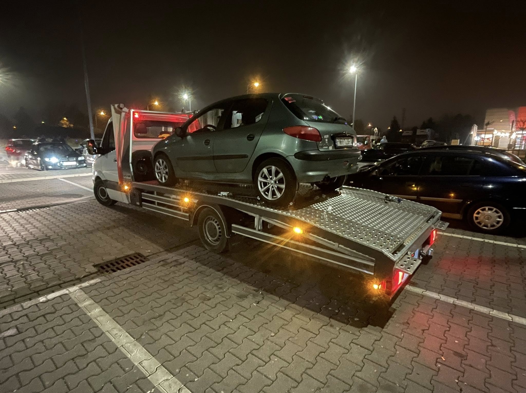 Laweta z zielonym Peugeot 206 na platformie, nocne oświetlenie uliczne, parking z kostki brukowej. Widoczne inne pojazdy i światła w tle.