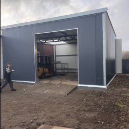 sebastian siegmuller - Nowoczesna hala magazynowa z szarej blachy w Lęborku, z otwartą bramą i wózkiem widłowym w środku. Osoba z notatnikiem wchodzi do środka. Widoczne elementy konstrukcyjne.