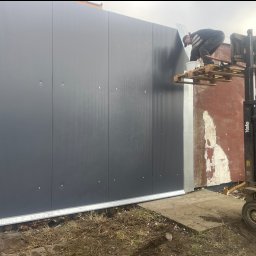 sebastian siegmuller - Montaż paneli ściennych na elewacji budynku z użyciem wózka widłowego jako platformy roboczej. Widoczny pracownik w trakcie pracy.