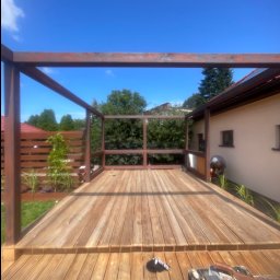 sebastian siegmuller - Drewniany taras z konstrukcją pergoli w trakcie budowy, widoczne deski, słupy i elementy mocujące. W tle zieleń ogrodu i fragment budynku.