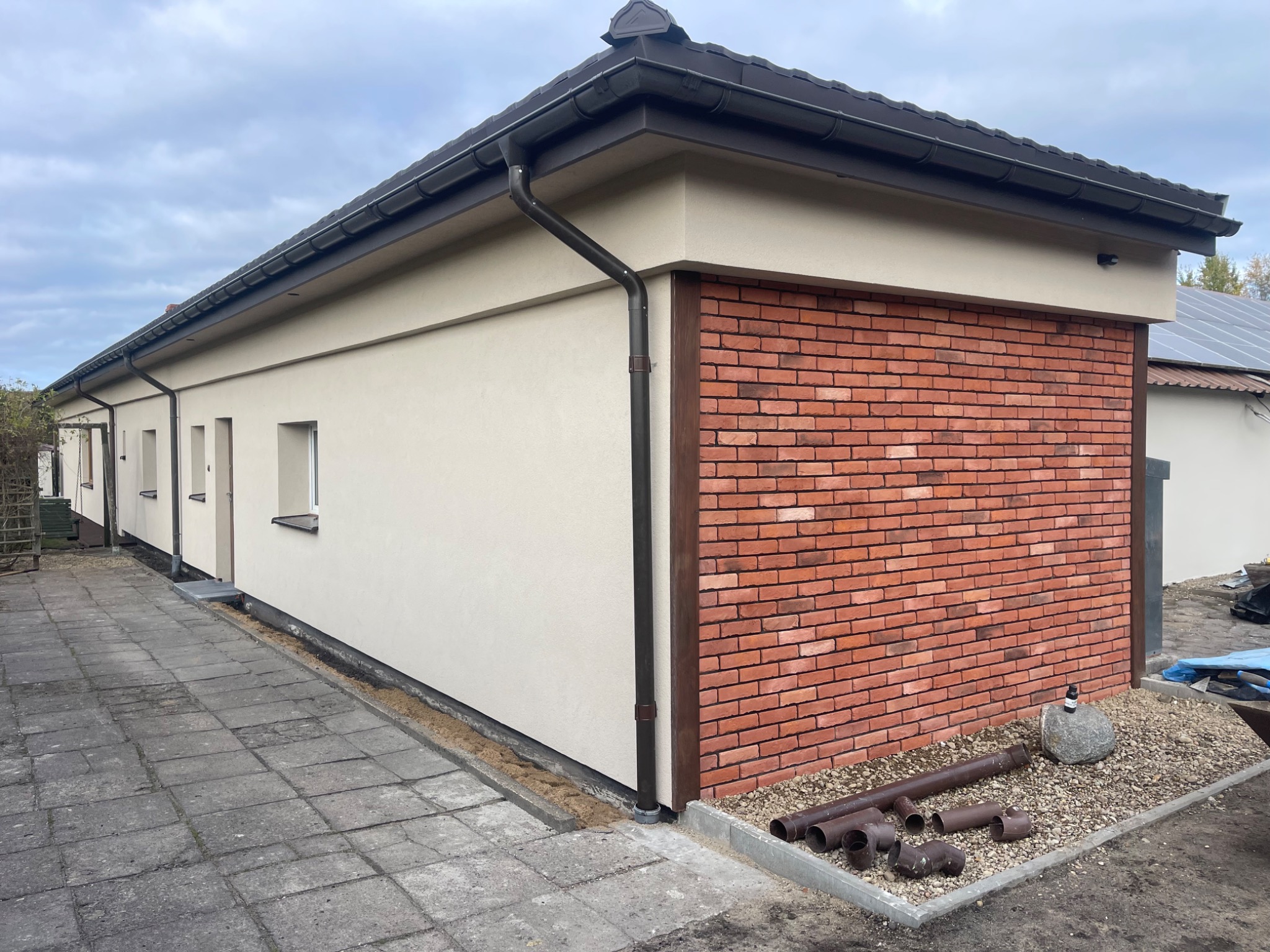 Wykończony budynek z elewacją w kolorze beżowym i fragmentem ściany z czerwonej cegły. Widoczne rynny, okno i chodnik z kostki brukowej. Na ziemi leżą rury.