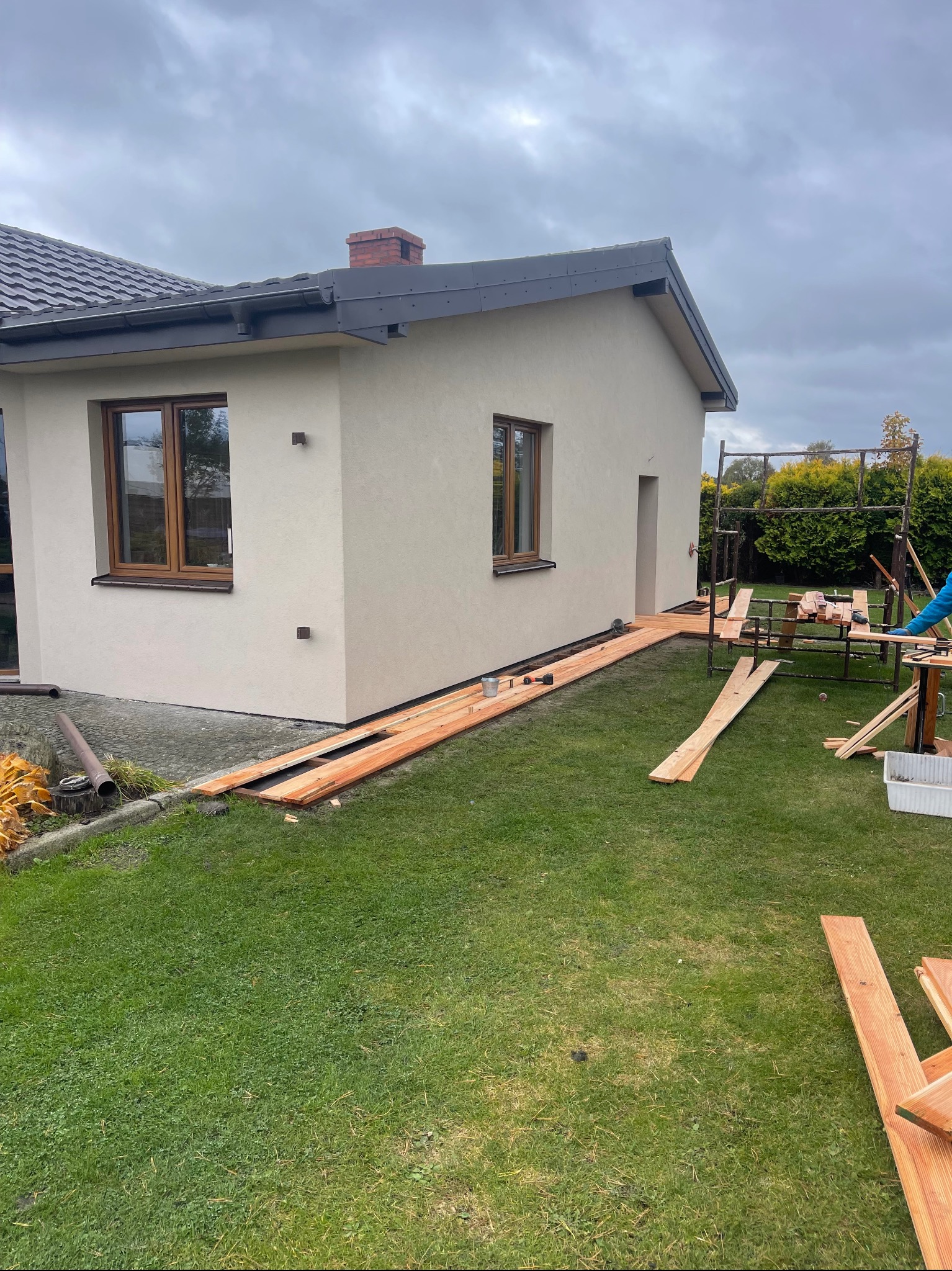 Montaż drewnianego tarasu przy domu jednorodzinnym. Widoczne deski tarasowe, narzędzia i fragment elewacji budynku z oknami. Prace w toku, widok z poziomu trawnika.