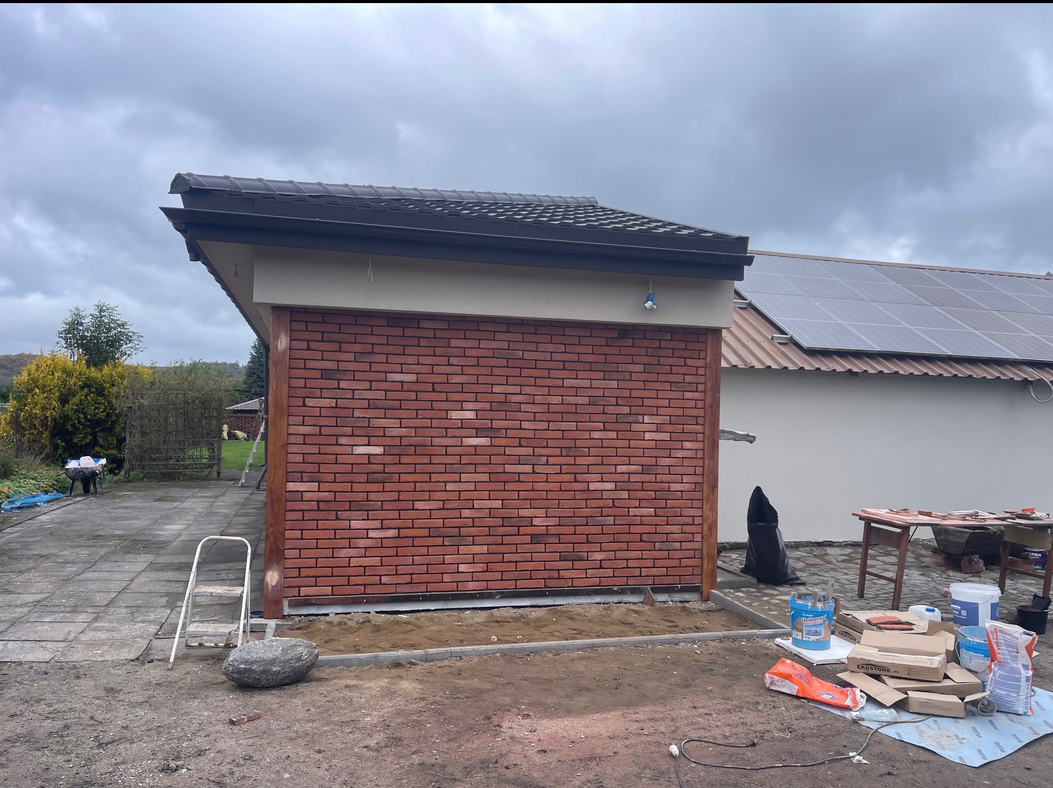 Ściana budynku wykończona czerwoną cegłą klinkierową, widoczne materiały budowlane i drabina. W tle budynek z panelami słonecznymi. Pochmurne niebo.