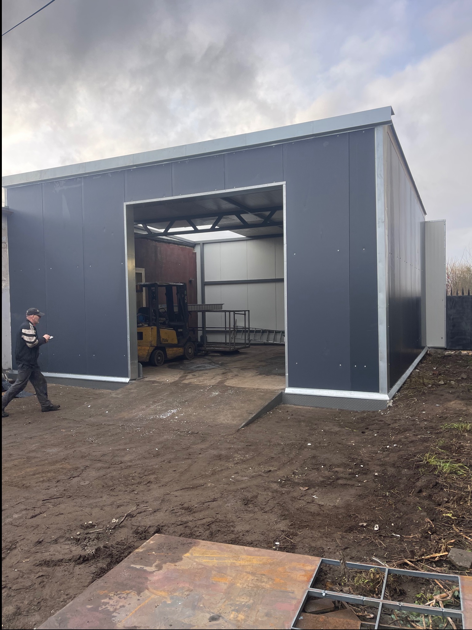 Nowoczesna hala magazynowa z szarej blachy w Lęborku, z otwartą bramą i wózkiem widłowym w środku. Osoba z notatnikiem wchodzi do środka. Widoczne elementy konstrukcyjne.