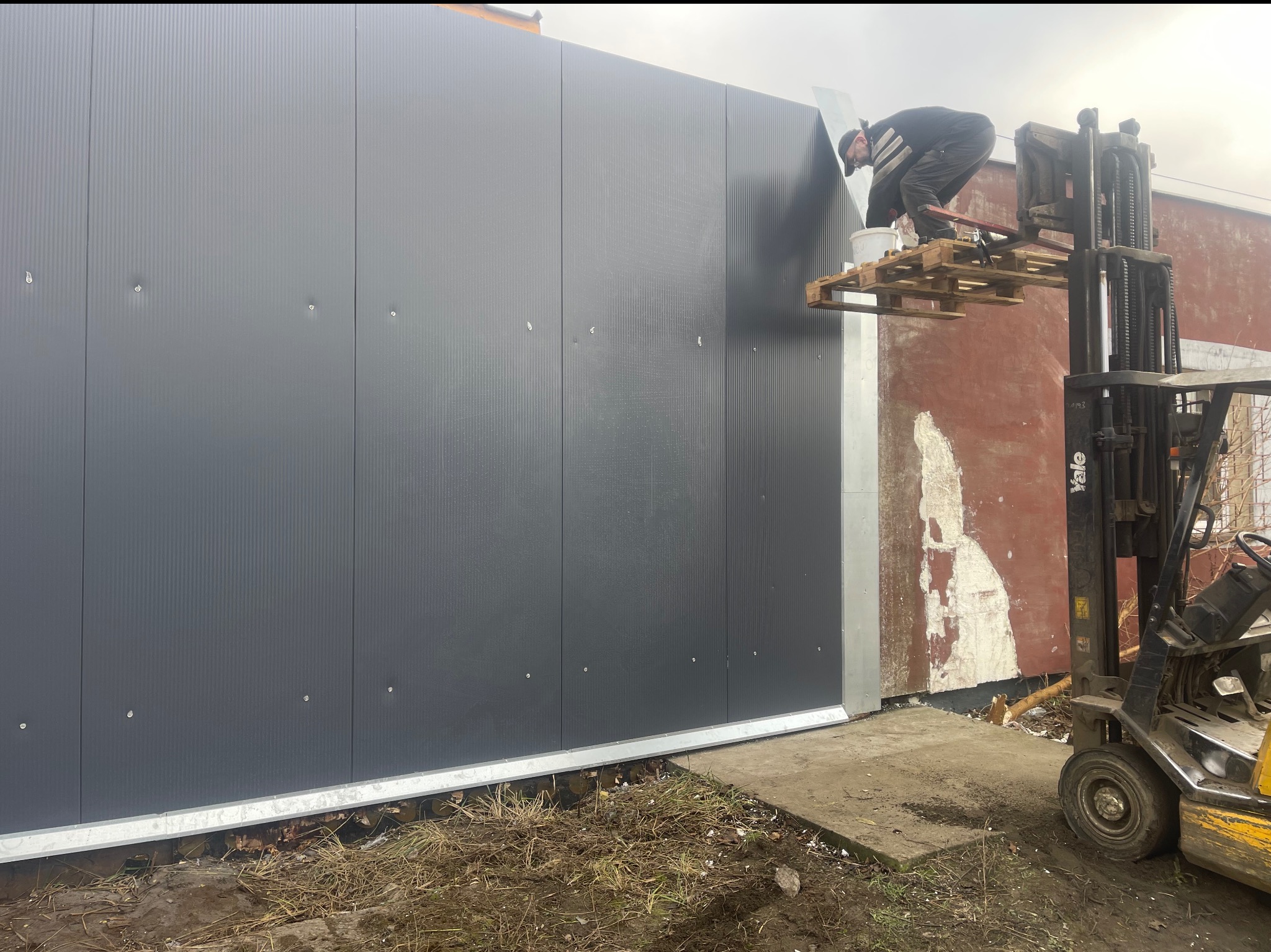 Montaż paneli ściennych na elewacji budynku z użyciem wózka widłowego jako platformy roboczej. Widoczny pracownik w trakcie pracy.