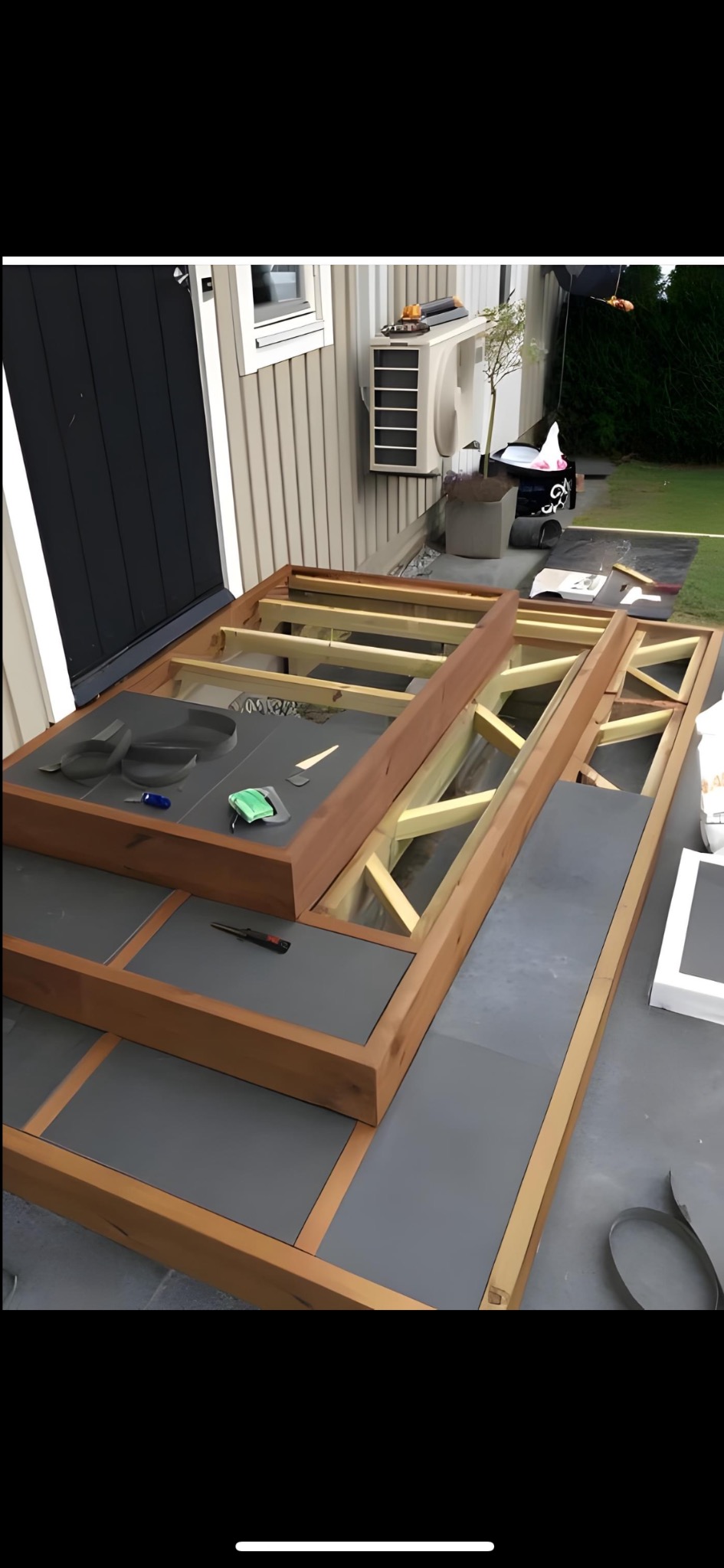 Drewniana konstrukcja tarasu w trakcie budowy, z szarymi płytami częściowo ułożonymi. Widoczne narzędzia i elementy montażowe. Tło stanowi elewacja budynku i zieleń.