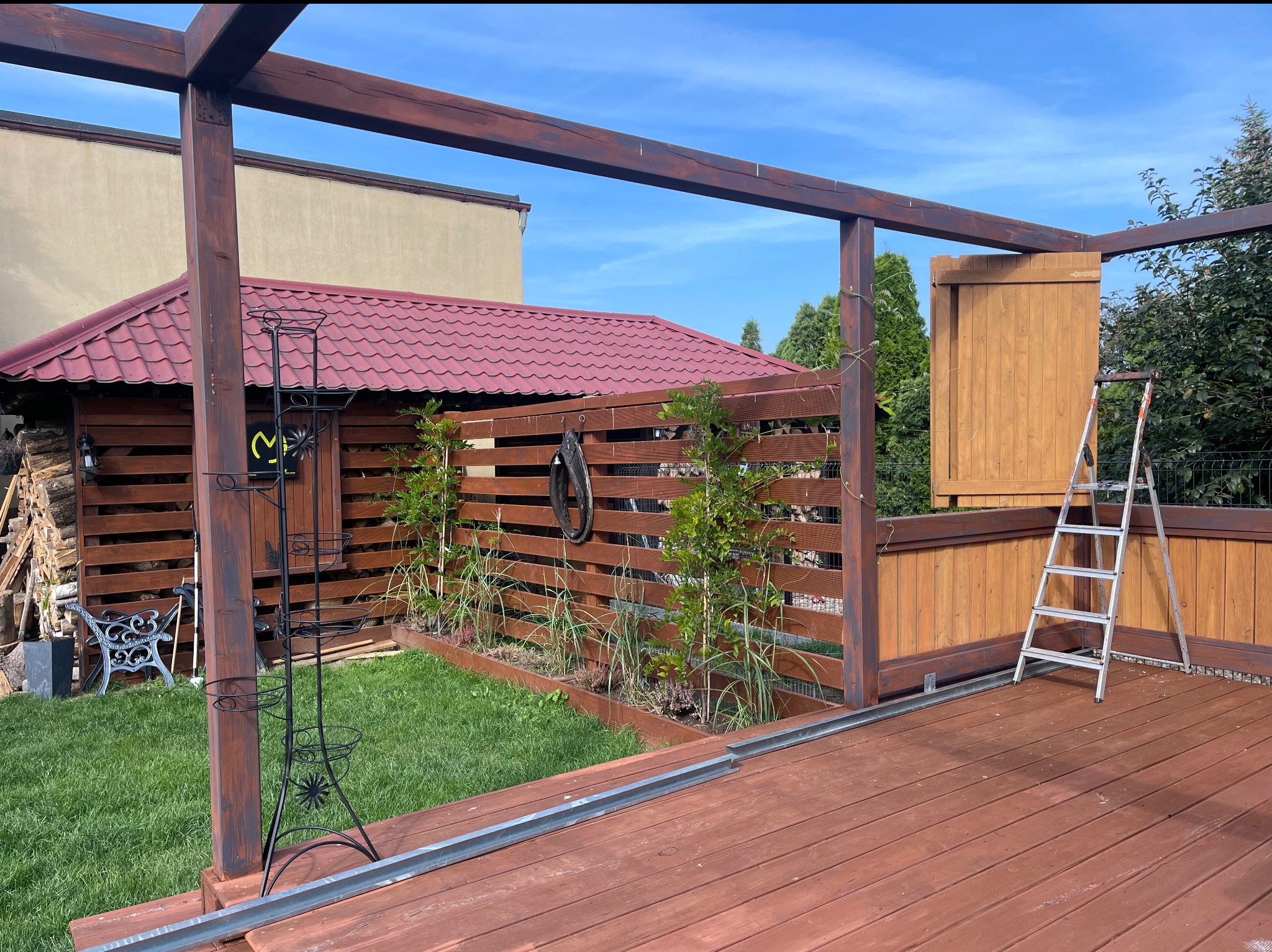 Drewniana pergola z zadaszeniem i elementami dekoracyjnymi, świeżo po impregnacji, z widoczną drabiną i metalowymi profilami na tarasie. Słoneczny dzień.