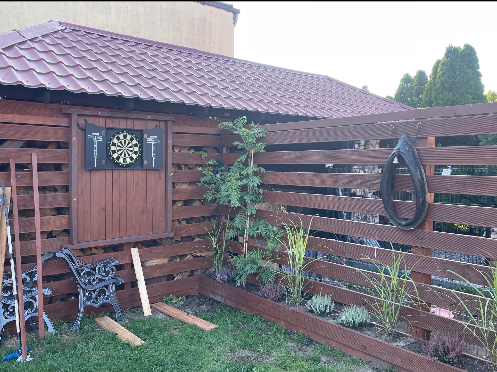 Drewniana altana z tarczą do darta, roślinami wzdłuż ogrodzenia i elementami dekoracyjnymi, takimi jak stare jarzmo. Widoczne drewno opałowe i metalowe elementy.