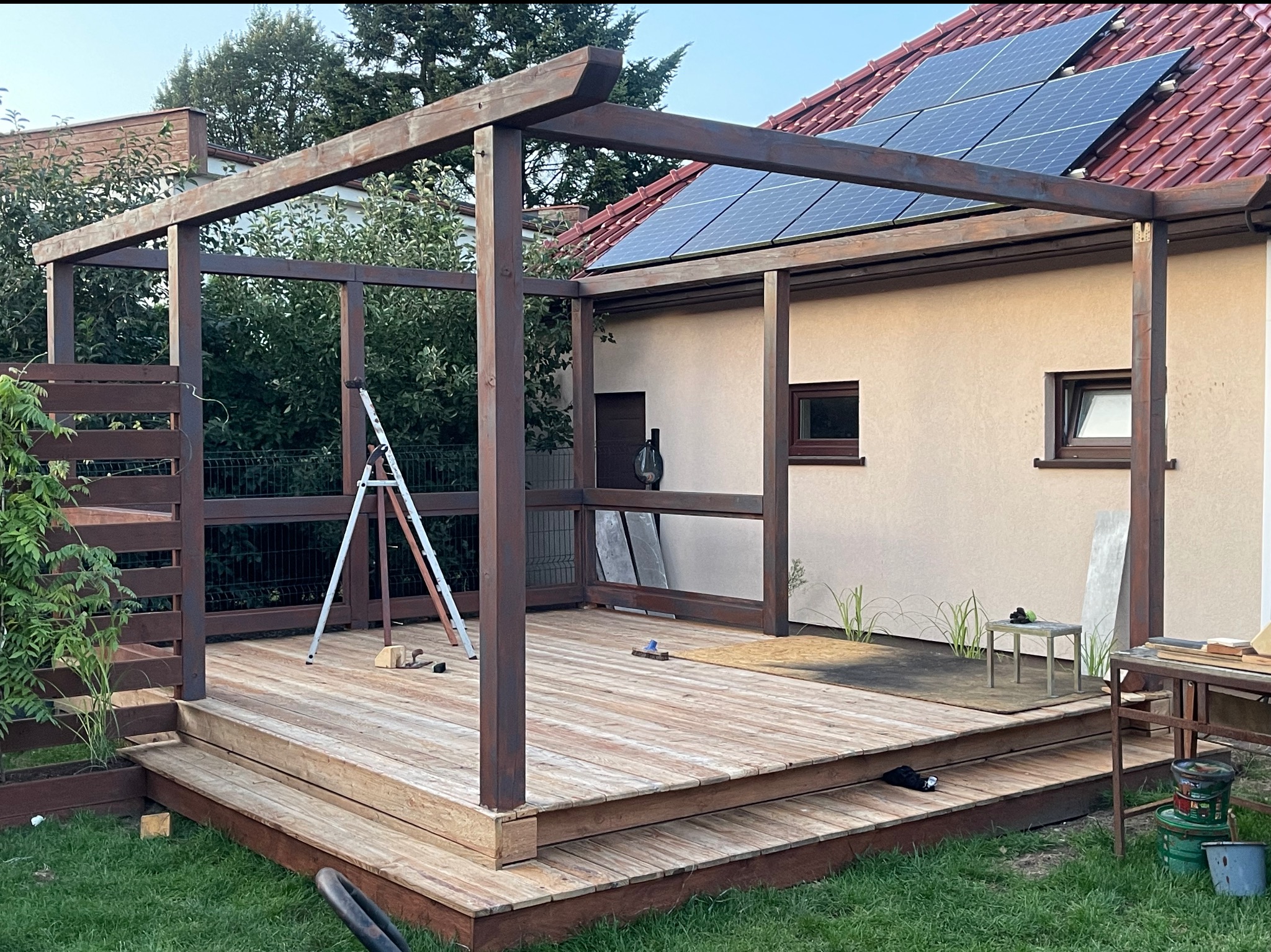 Drewniana pergola z tarasem w ogrodzie, widoczne panele słoneczne na dachu domu. Elementy konstrukcyjne pomalowane na ciemny brąz, schody prowadzące na taras.