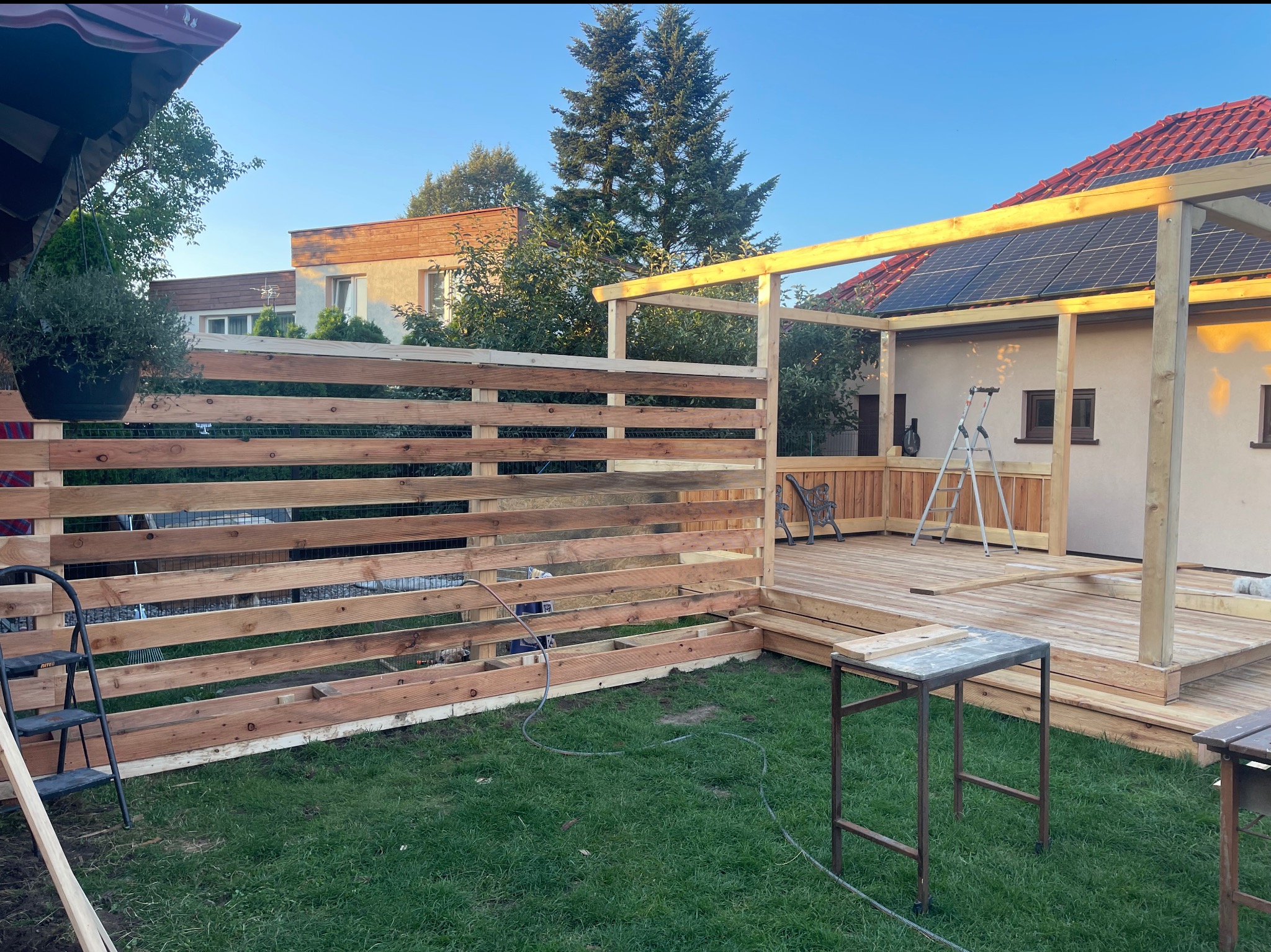 Drewniana konstrukcja tarasu z ażurowym ogrodzeniem w trakcie budowy, widoczna drabina i narzędzia, trawnik w tle, dom z panelami słonecznymi.