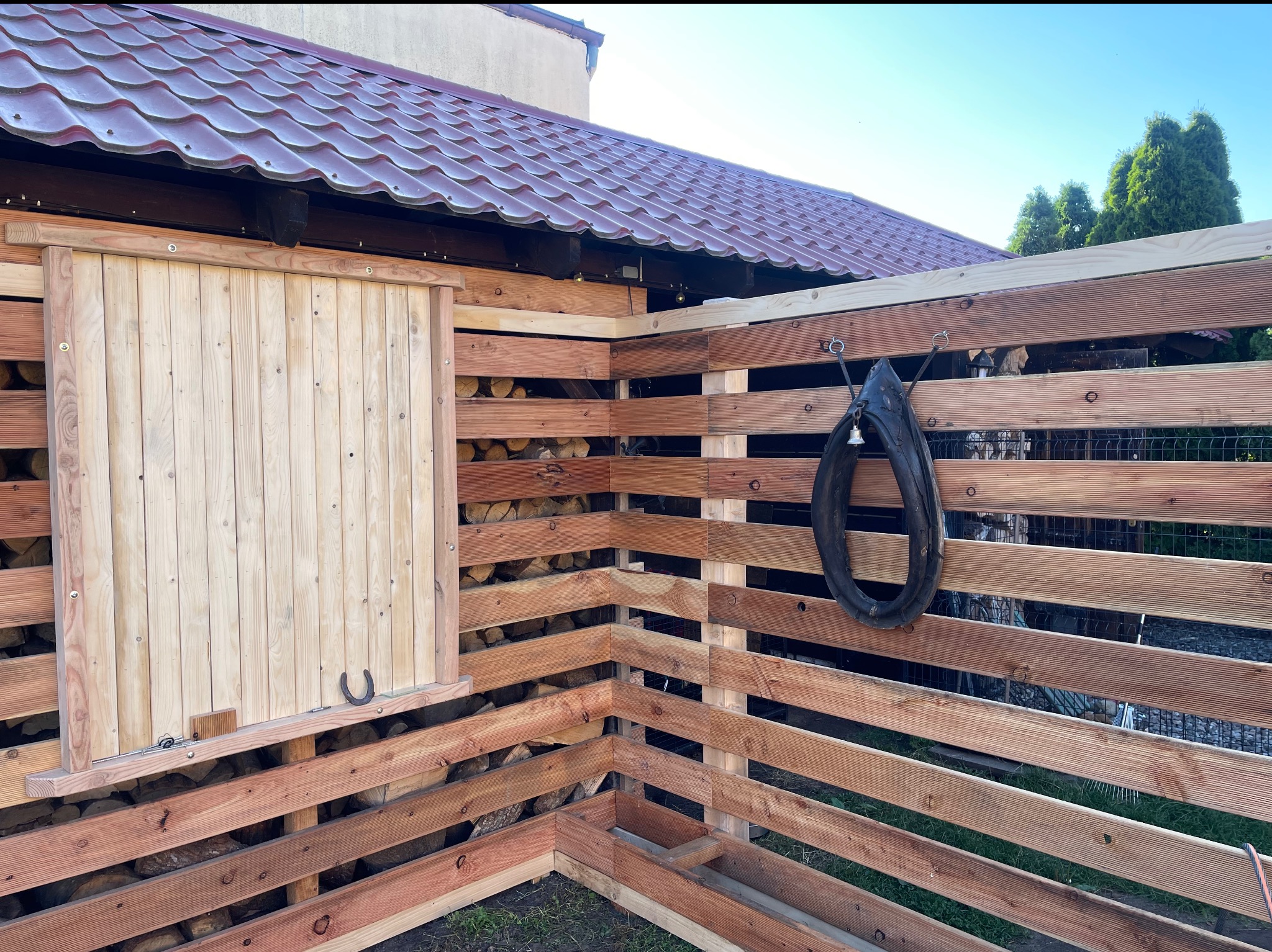 Drewniana konstrukcja z przesuwnymi drzwiami, wypełniona pociętym drewnem. Na zewnątrz wisi stara opona. Widoczny fragment dachu i zieleni.