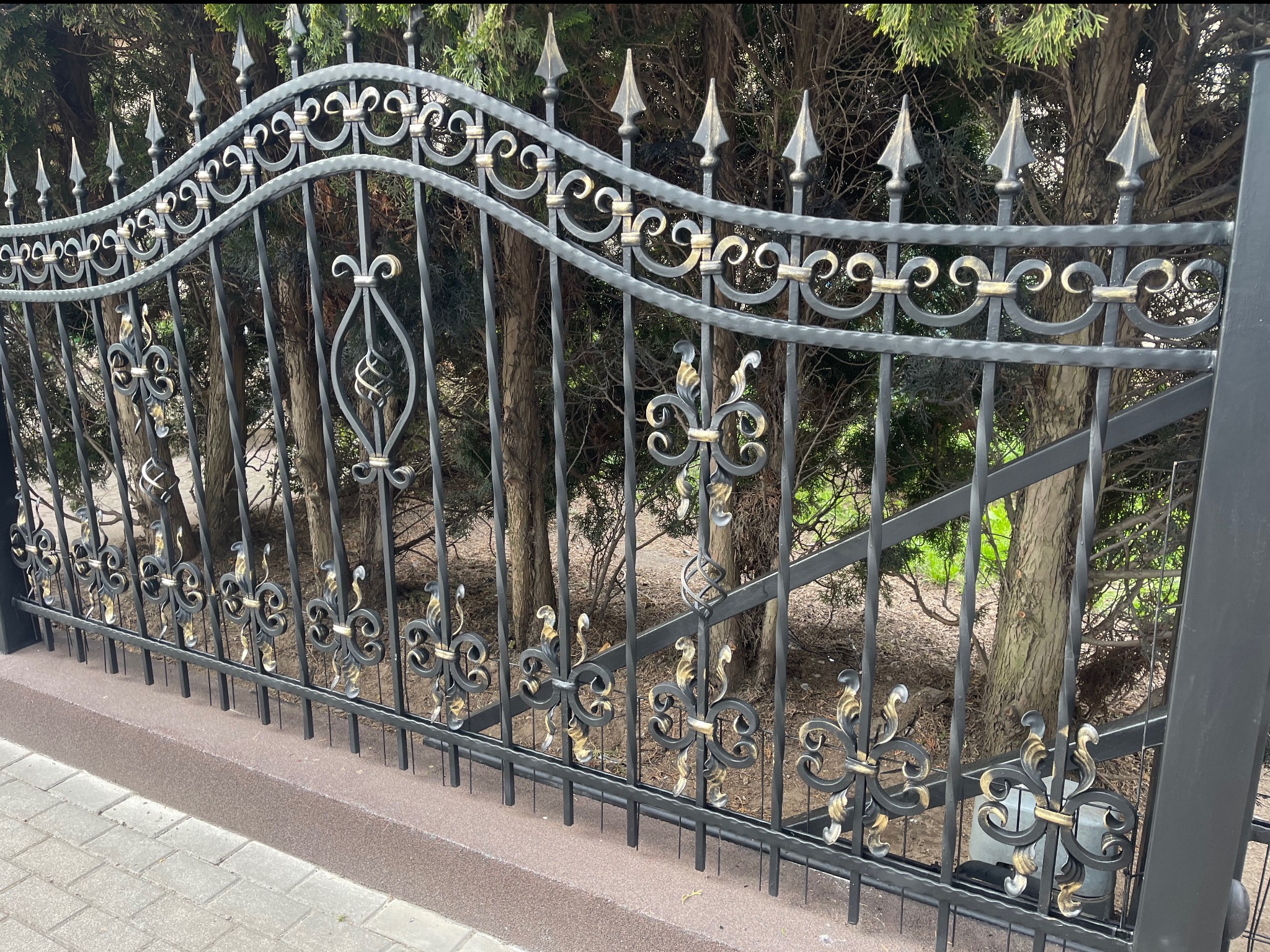 Kuta, dekoracyjna brama w kolorze grafitowym z elementami złota na tle zielonych drzew. Widoczny fragment chodnika z kostki brukowej.