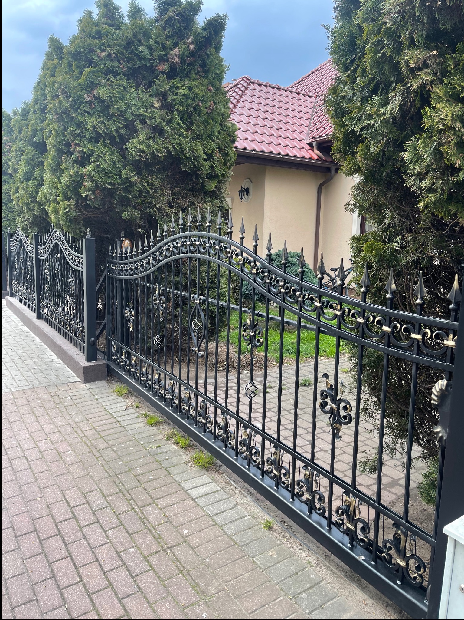 Czarna, metalowa brama z ozdobnymi elementami w złotym kolorze, prowadząca do posesji z czerwonym dachem, otoczonej tujami i brukowanym chodnikiem.