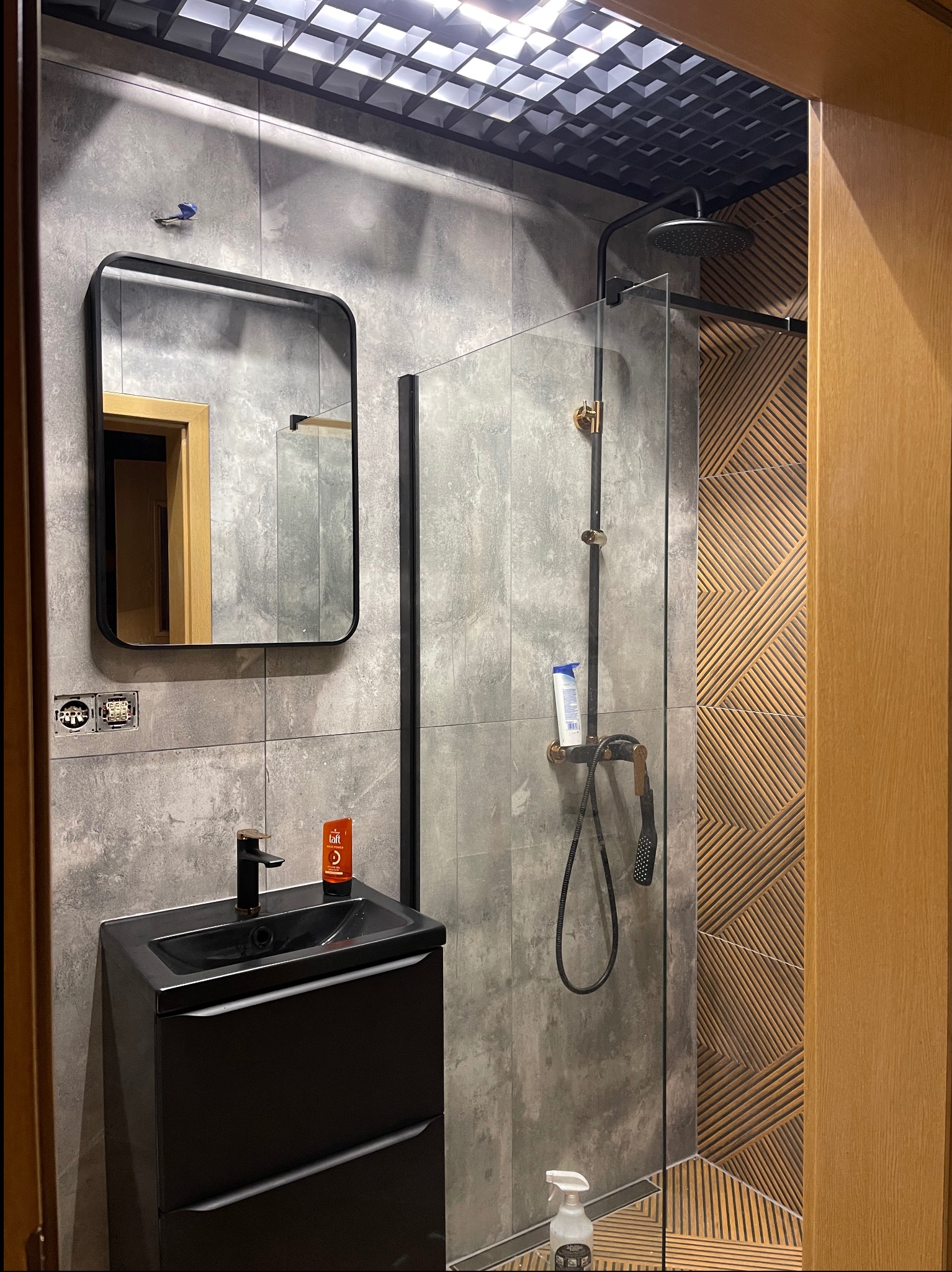 Nowoczesna łazienka z czarną armaturą, szarymi płytkami imitującymi beton i dekoracyjną ścianą z drewnopodobnych paneli. Kabina prysznicowa ze szklaną ścianką.