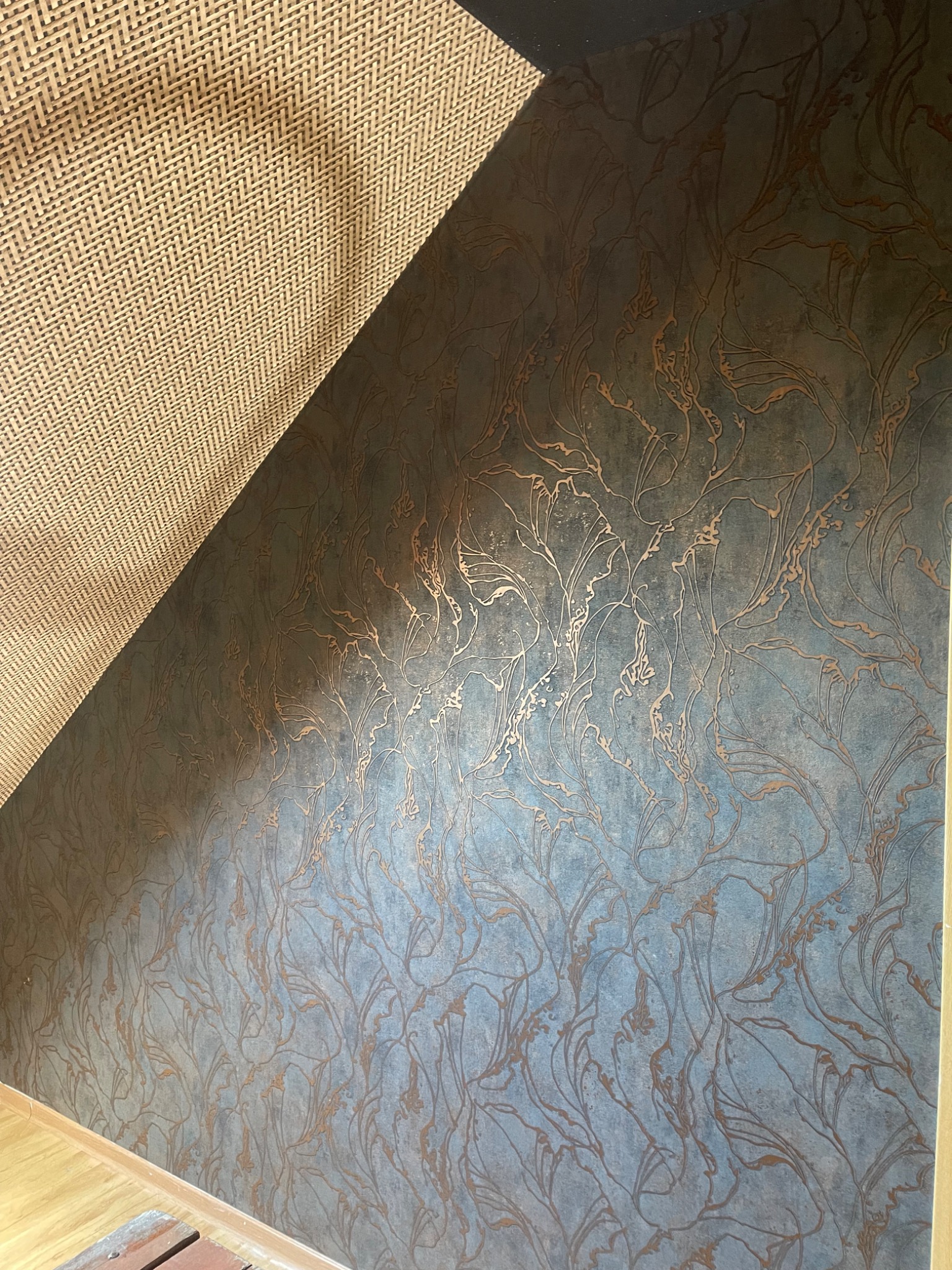 Ściana w pokoju na poddaszu, pokryta tapetą w odcieniach szarości i brązu z motywem roślinnym. Widoczny fragment sufitu z wykończeniem w jodełkę.